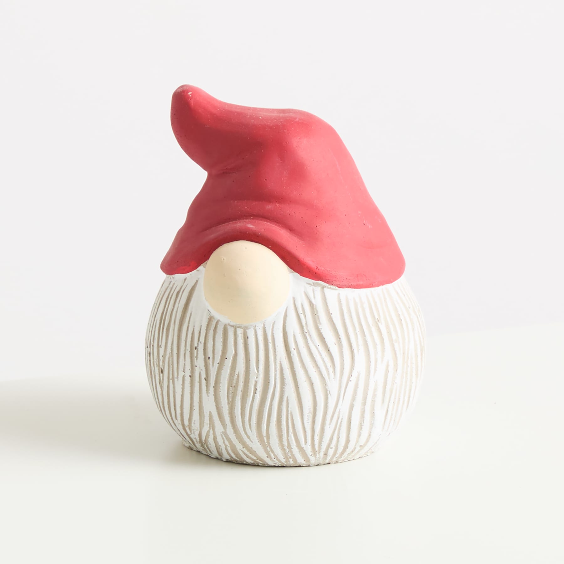 Eine kleine, runde Keramikfigur mit rotem Hut und weißem, strukturiertem Körper, die einem Gnom oder einer Gartenfigur ähnelt, sitzt auf einer schlichten, hellen Oberfläche.