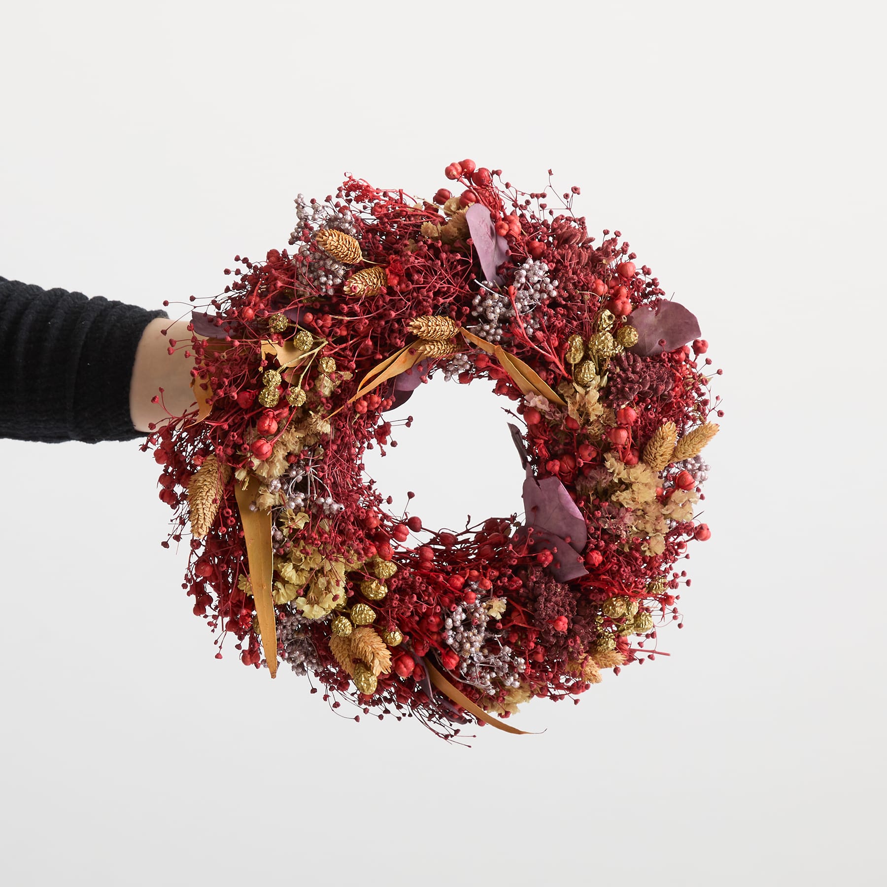 Ein farbenfroher, runder Kranz aus verschiedenen getrockneten Blumen, Beeren und anderen natürlichen Elementen wird vor einem schlichten weißen Hintergrund in den Händen einer Person gehalten.
