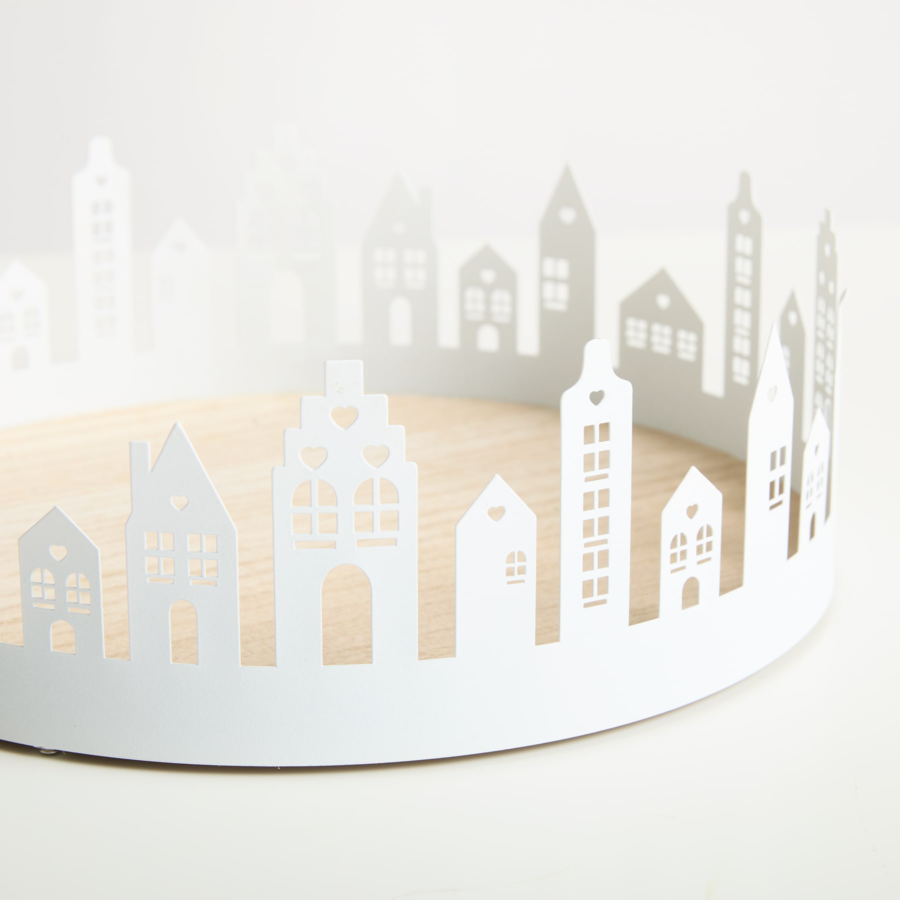 Ein weißer, runder Tortenständer mit einem ausgeschnittenen Design, das vor einem schlichten weißen Untergrund eine Stadtlandschaft mit verschiedenen Gebäuden und Strukturen im Hintergrund zeigt.