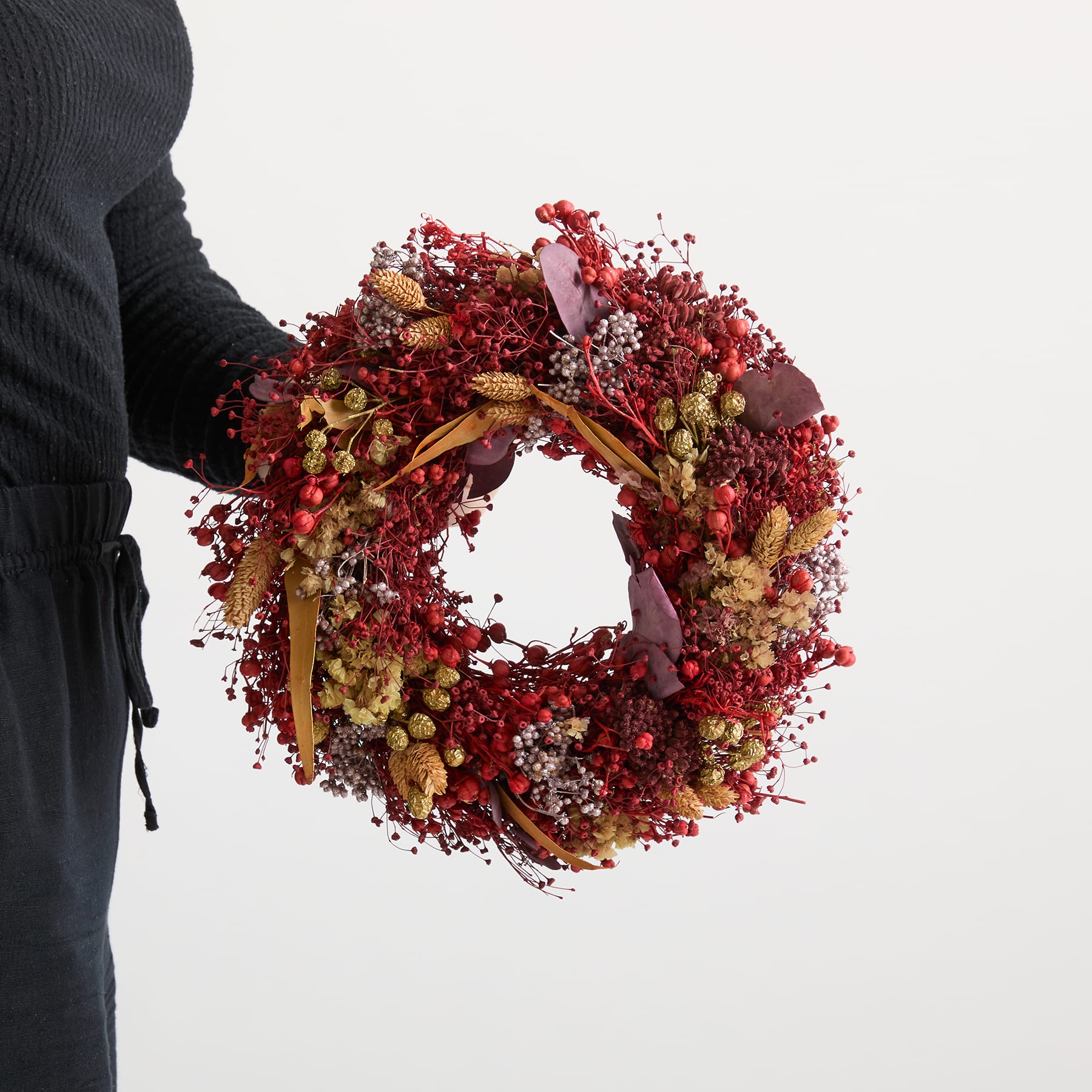 Ein lebendiger, festlicher Kranz aus verschiedenen roten und goldenen Beeren, Blumen und anderen natürlichen Elementen wird im Vordergrund vor einem schlichten weißen Hintergrund gehalten.