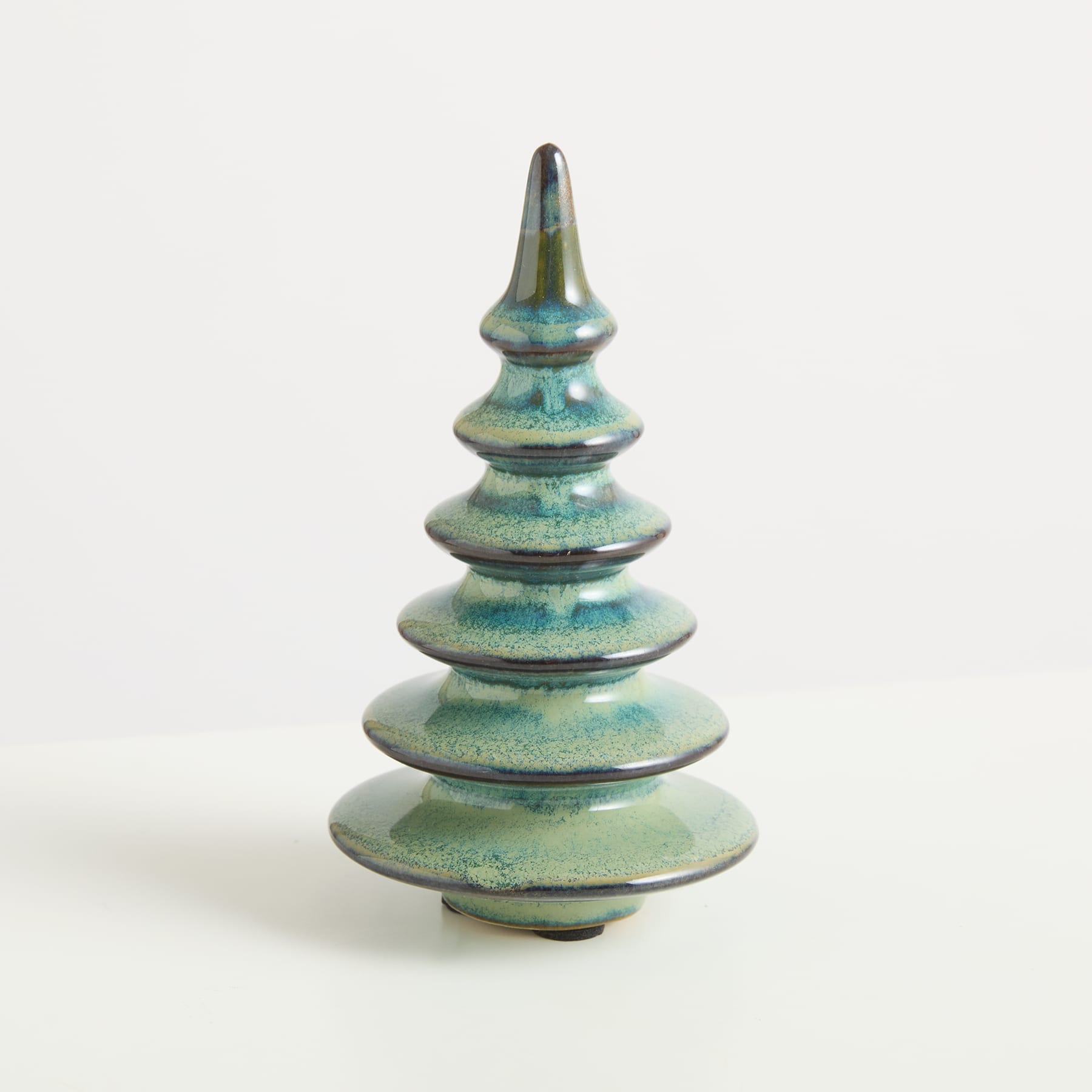 Eine Keramikskulptur, die einem stilisierten Weihnachtsbaum ähnelt und eine grüne und blaue Glasur aufweist, steht vor einem schlichten weißen Hintergrund.