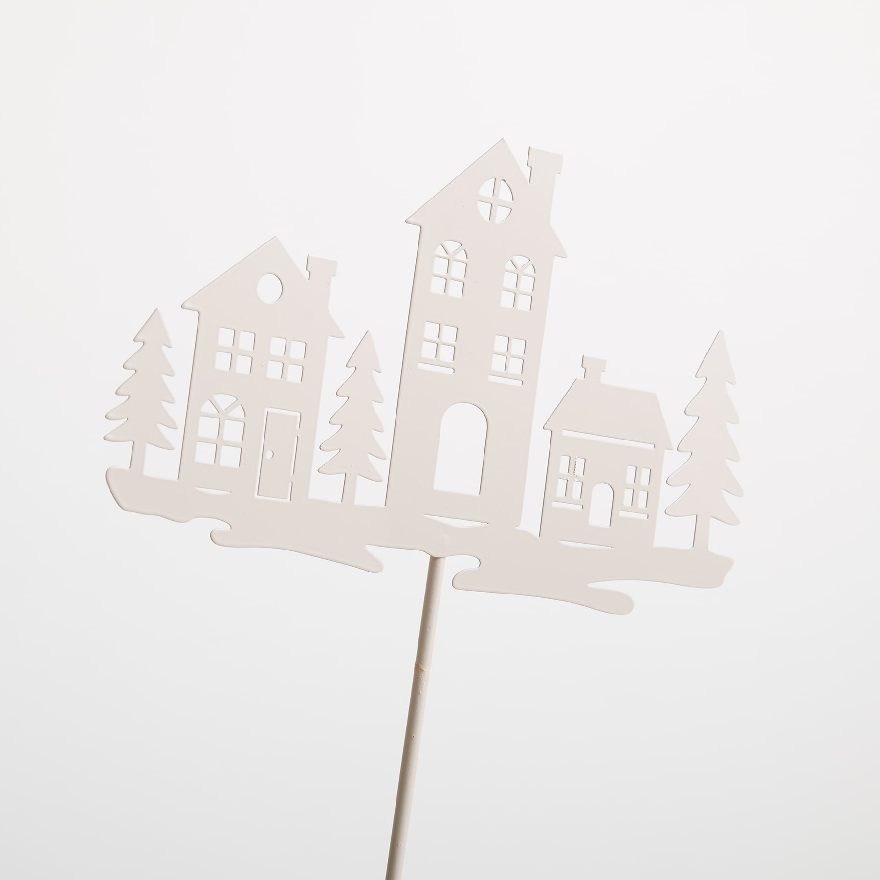 Ein weißer Papierausschnitt, der eine Winterlandschaft mit Häusern, Bäumen und einem Kirchturm darstellt, montiert auf einem Holzstab.