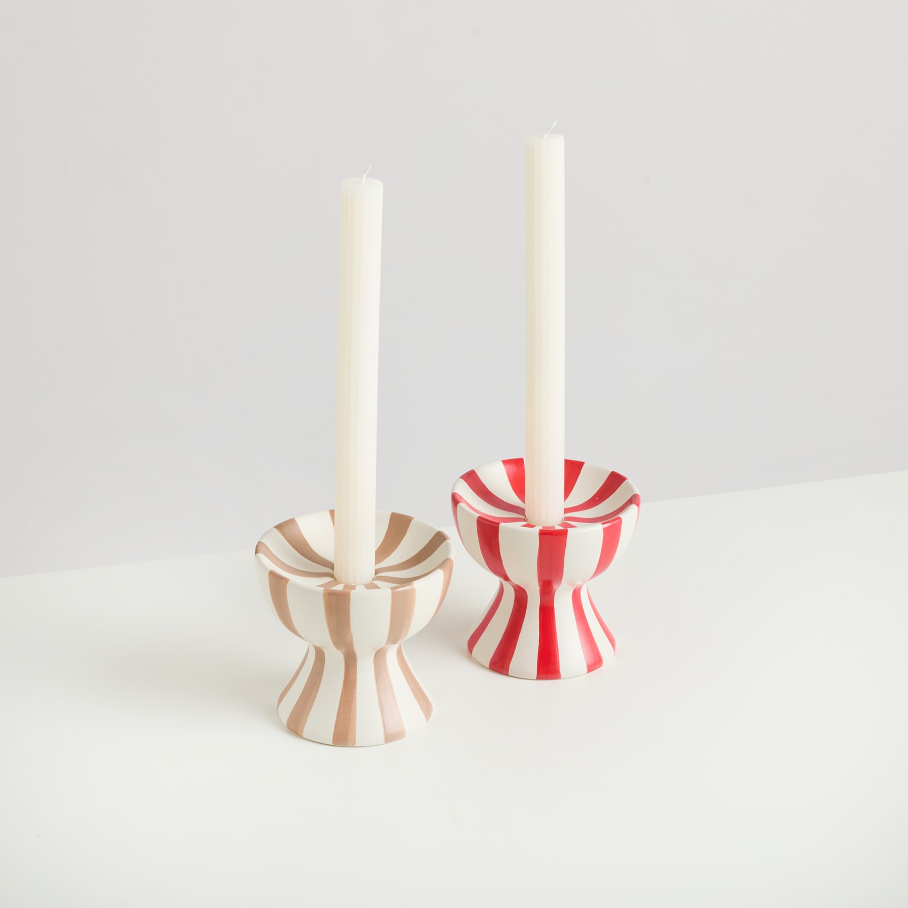 Zwei Kerzenständer aus Holz, einer mit rot-weißem Streifenmuster und der andere mit schlichtem weißem Design, stehen auf einer hellen Fläche vor einem schlichten Hintergrund.