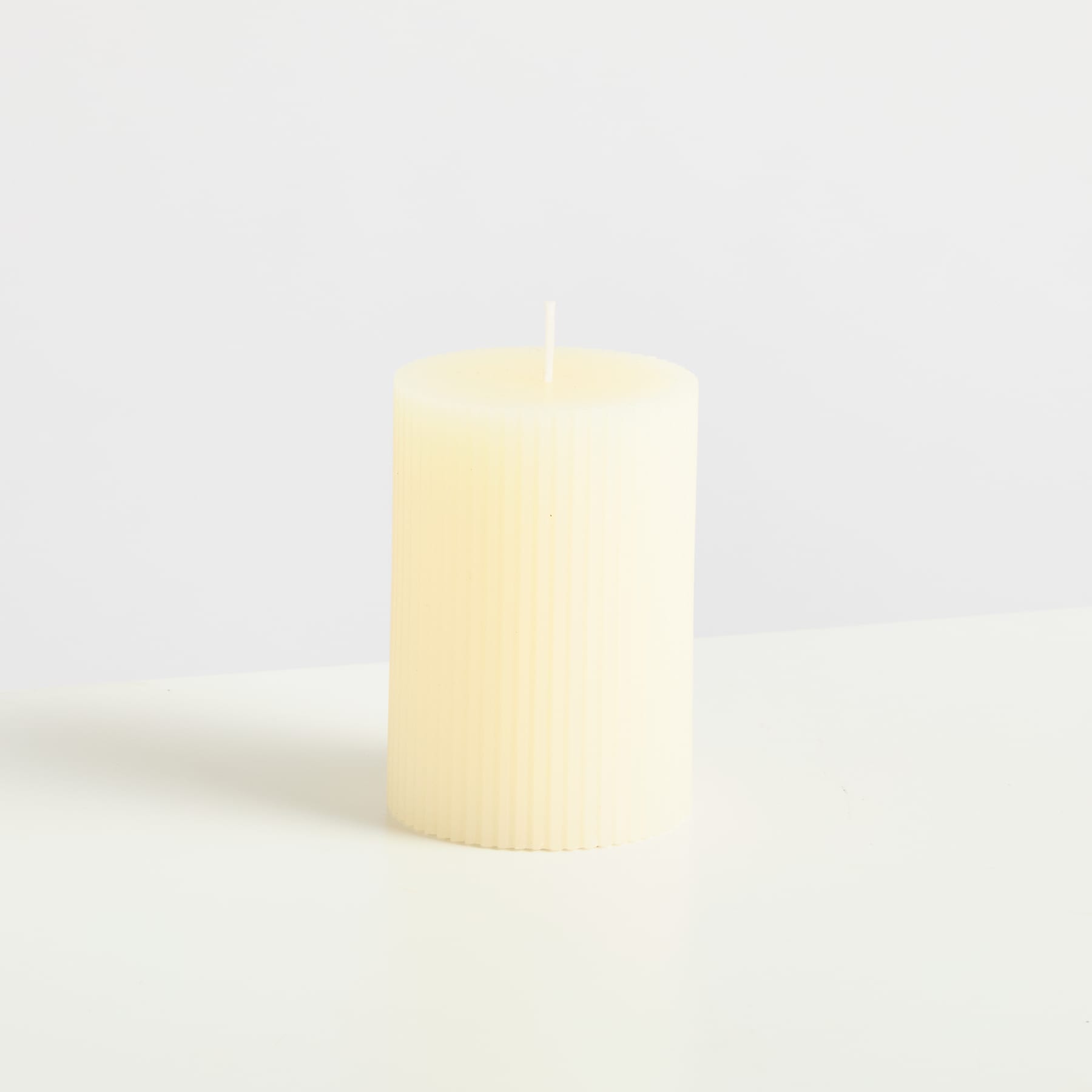 Eine einfache, zylindrische Kerze mit einem weichen, warmen Schein steht allein auf einer schlichten, hellen Oberfläche.