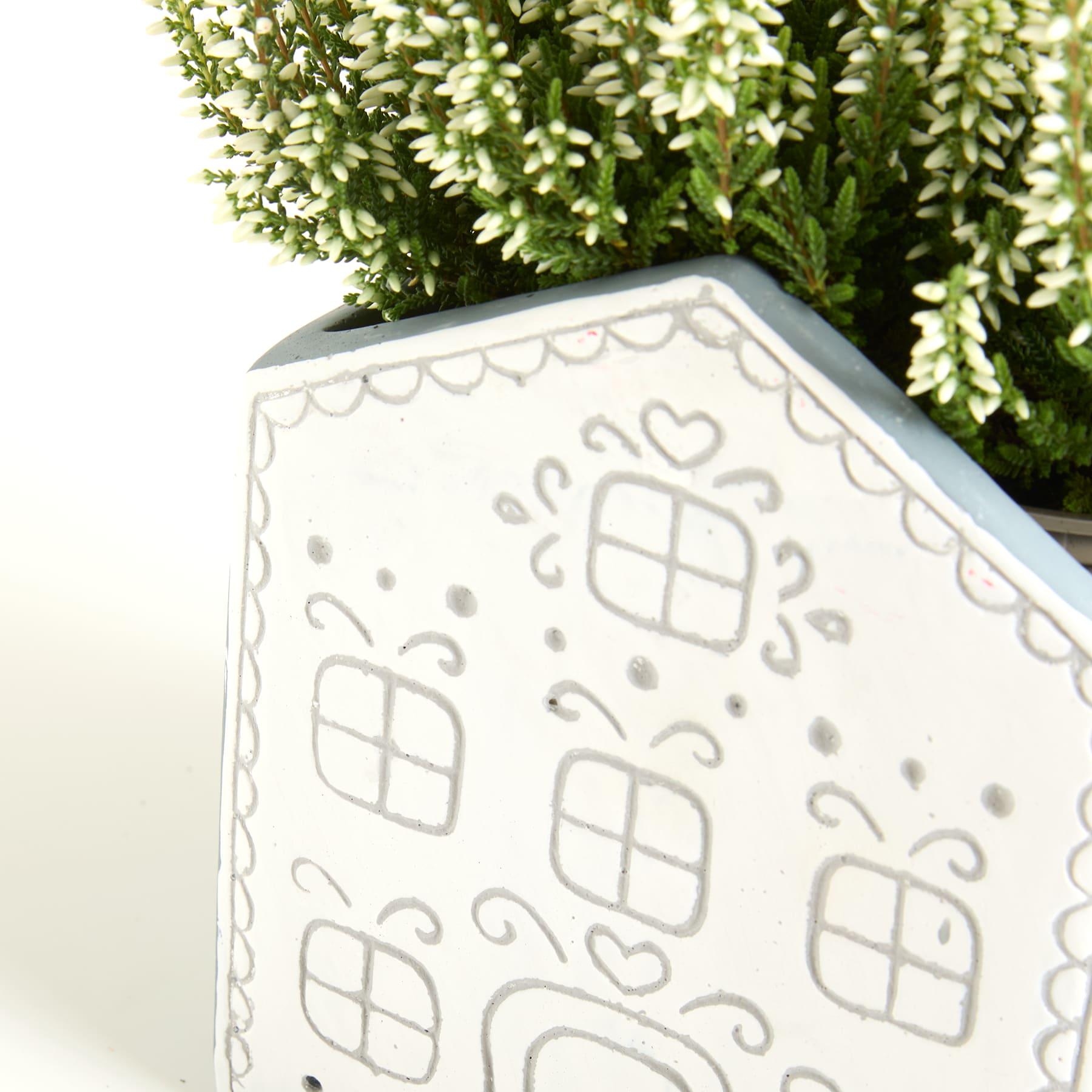 Ein weißer Keramiktopf mit einem dekorativen Muster aus geometrischen Formen steht vor einer üppigen, grünen Pflanze.