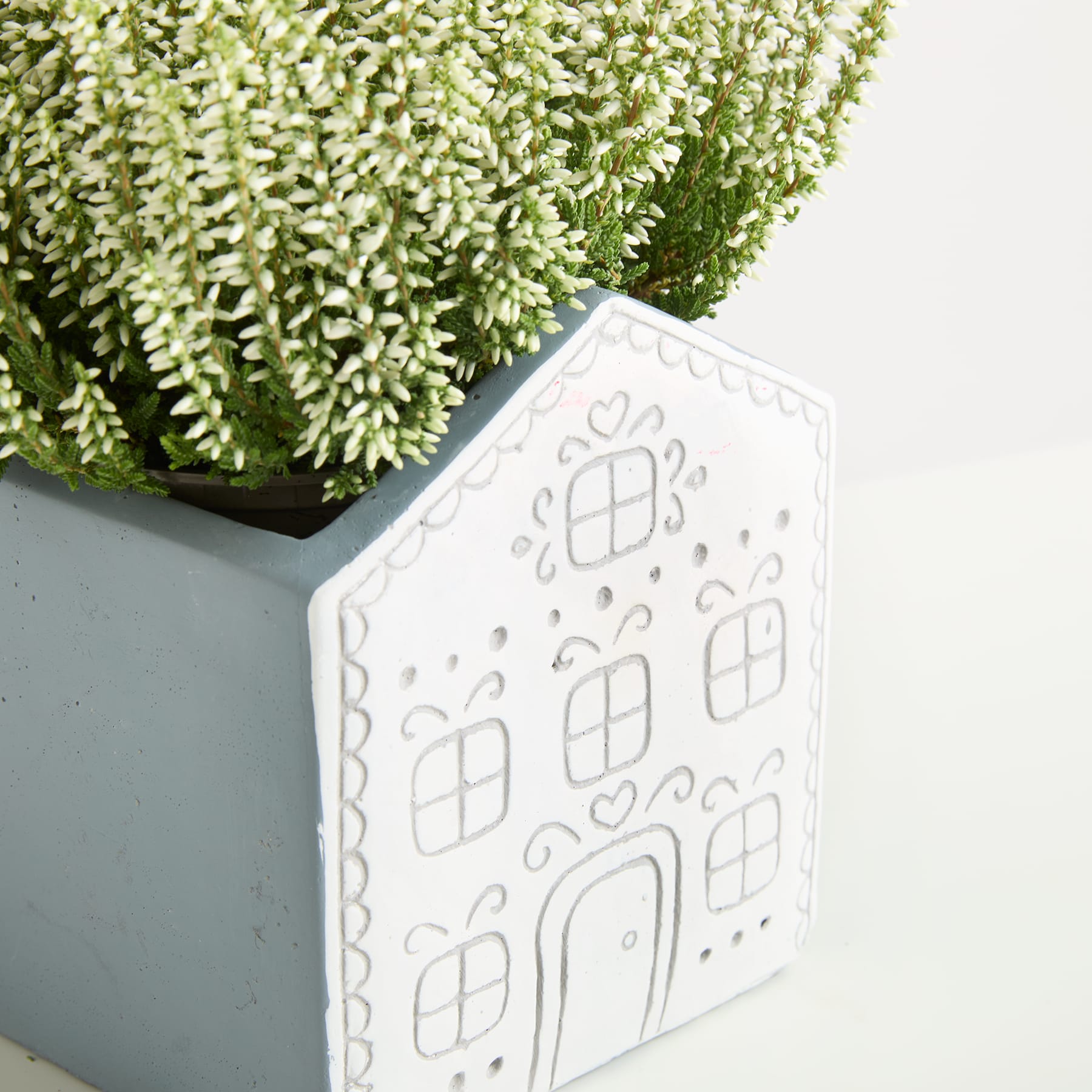 Ein weißes, dekoratives Objekt in Hausform mit komplizierten Mustern steht vor einer üppigen, grünen Pflanze.
