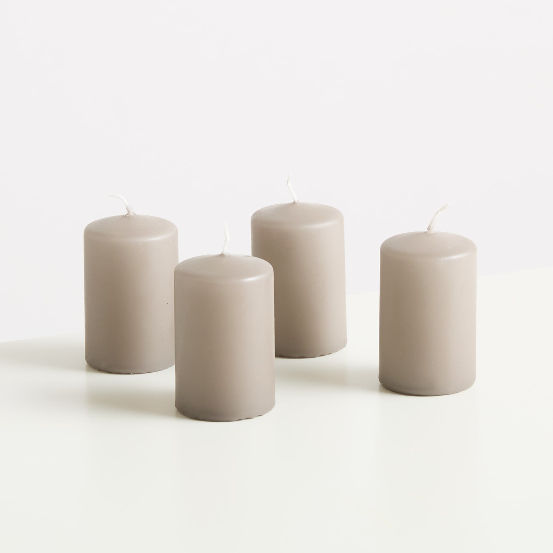 Vier einfache, zylindrische Kerzen in einem neutralen, cremefarbenen Farbton stehen auf einer schlichten, hellen Oberfläche vor einem schlichten, weißen Hintergrund.