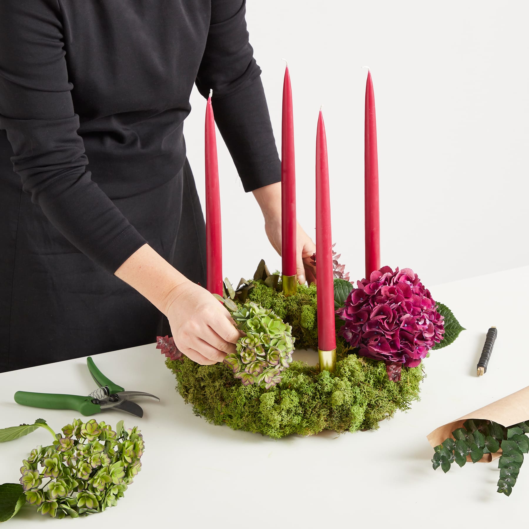 Das Bild zeigt die Hände einer Person, die ein Blumengesteck mit roten Kerzen, grünem Moos und rosa Blumen auf einer weißen Oberfläche arrangiert.