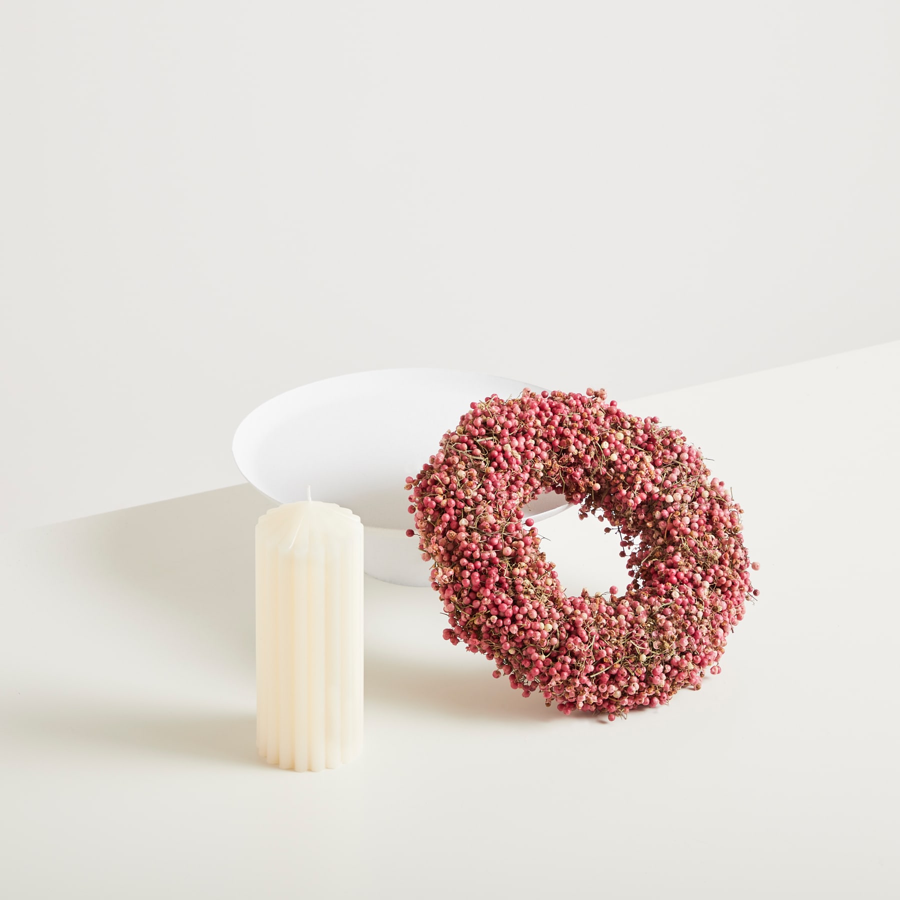 Vor einem schlichten weißen Hintergrund steht ein weißes zylindrisches Objekt neben einer kranzartigen Struktur, die mit kleinen rosa und roten Beeren oder Samen bedeckt ist.