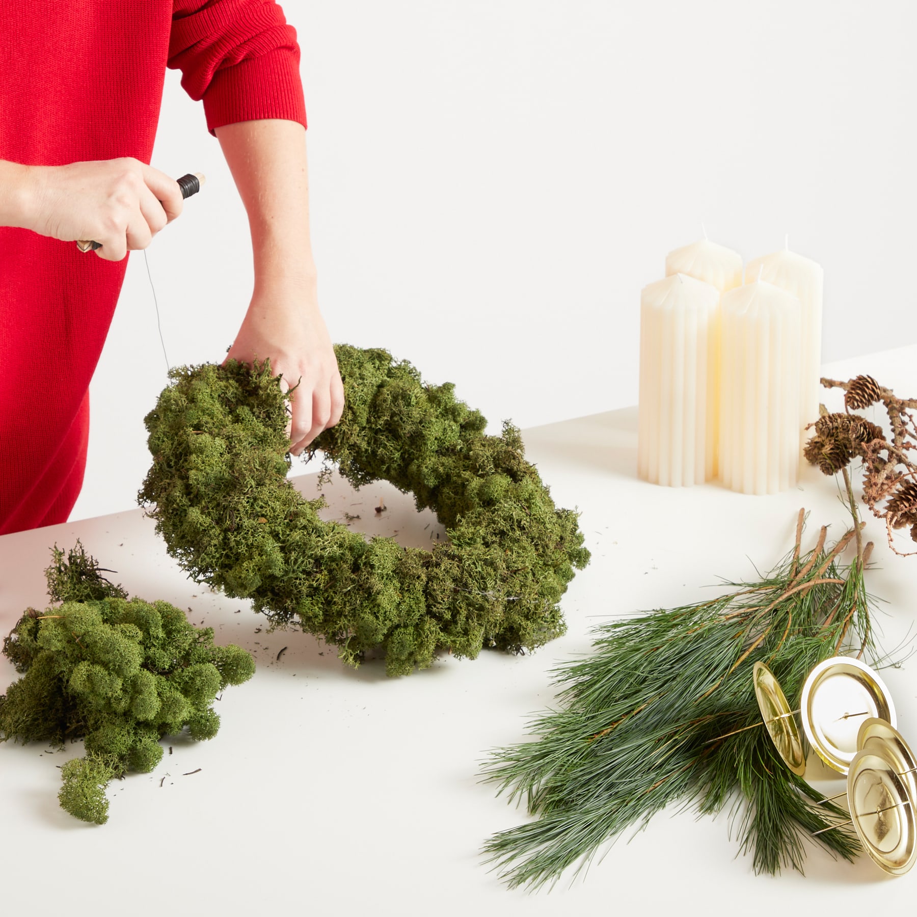 Das Bild zeigt die Hände einer Person, die einen grünen Mooskranz auf einer weißen Oberfläche arrangiert, mit Tannenzweigen und Kerzen im Hintergrund.