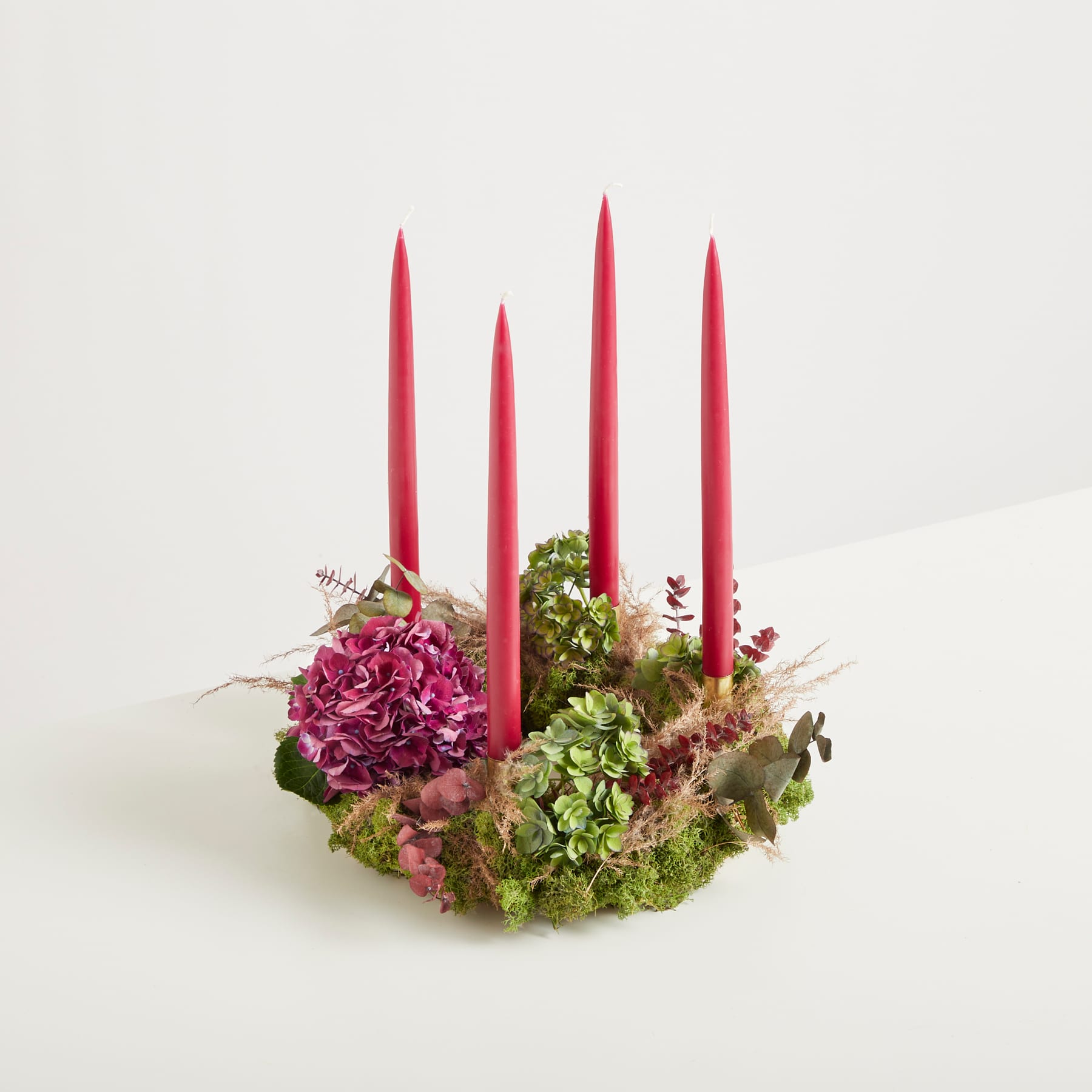 Auf einem schlichten weißen Hintergrund befindet sich ein kreisförmiges Arrangement leuchtend roter Kerzen, umgeben von üppigen, farbenfrohen Blumen und Grünpflanzen.