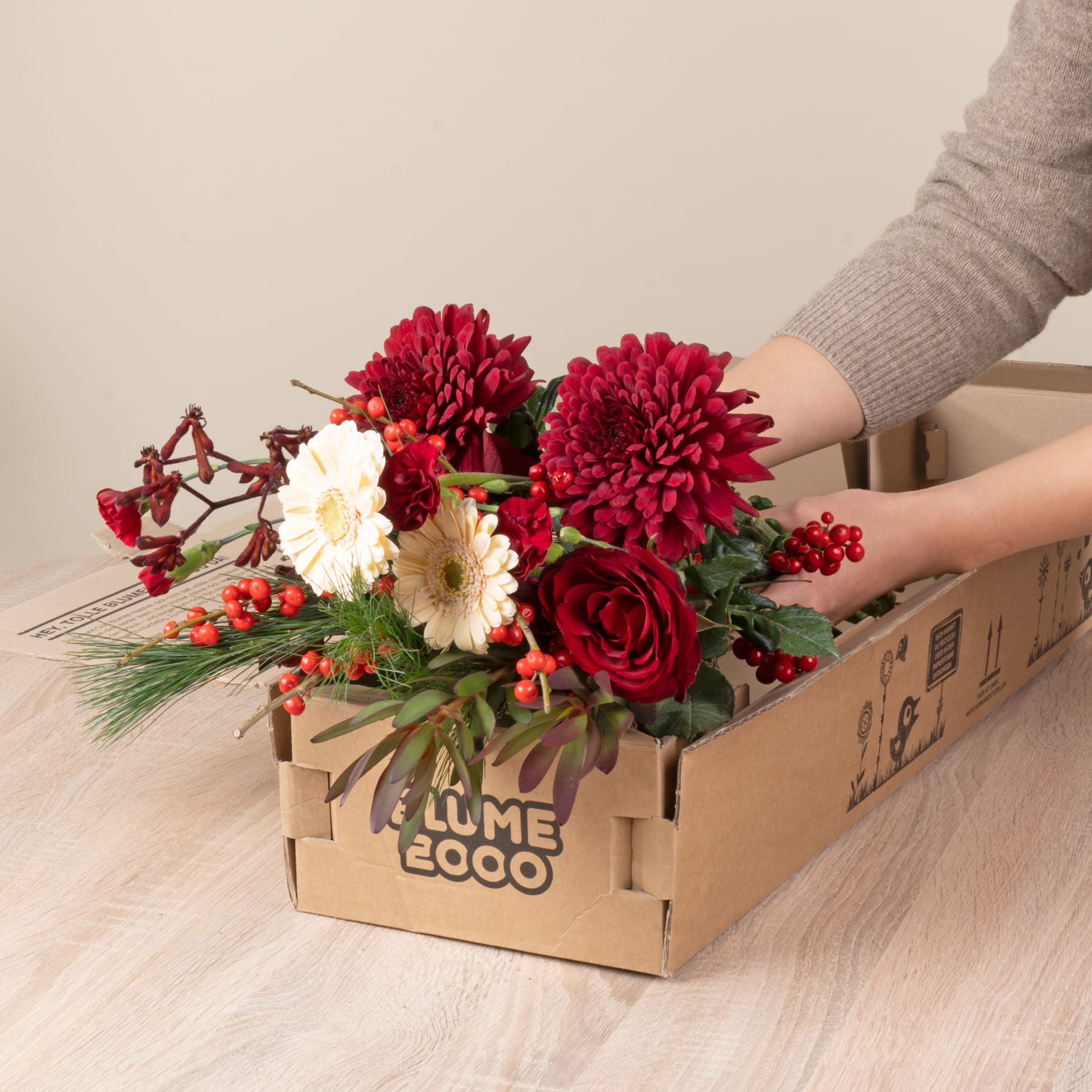 Auf einer Holzoberfläche steht eine Holzkiste mit einem farbenfrohen Strauß roter und weißer Blumen, darunter Rosen, Chrysanthemen und Beeren.