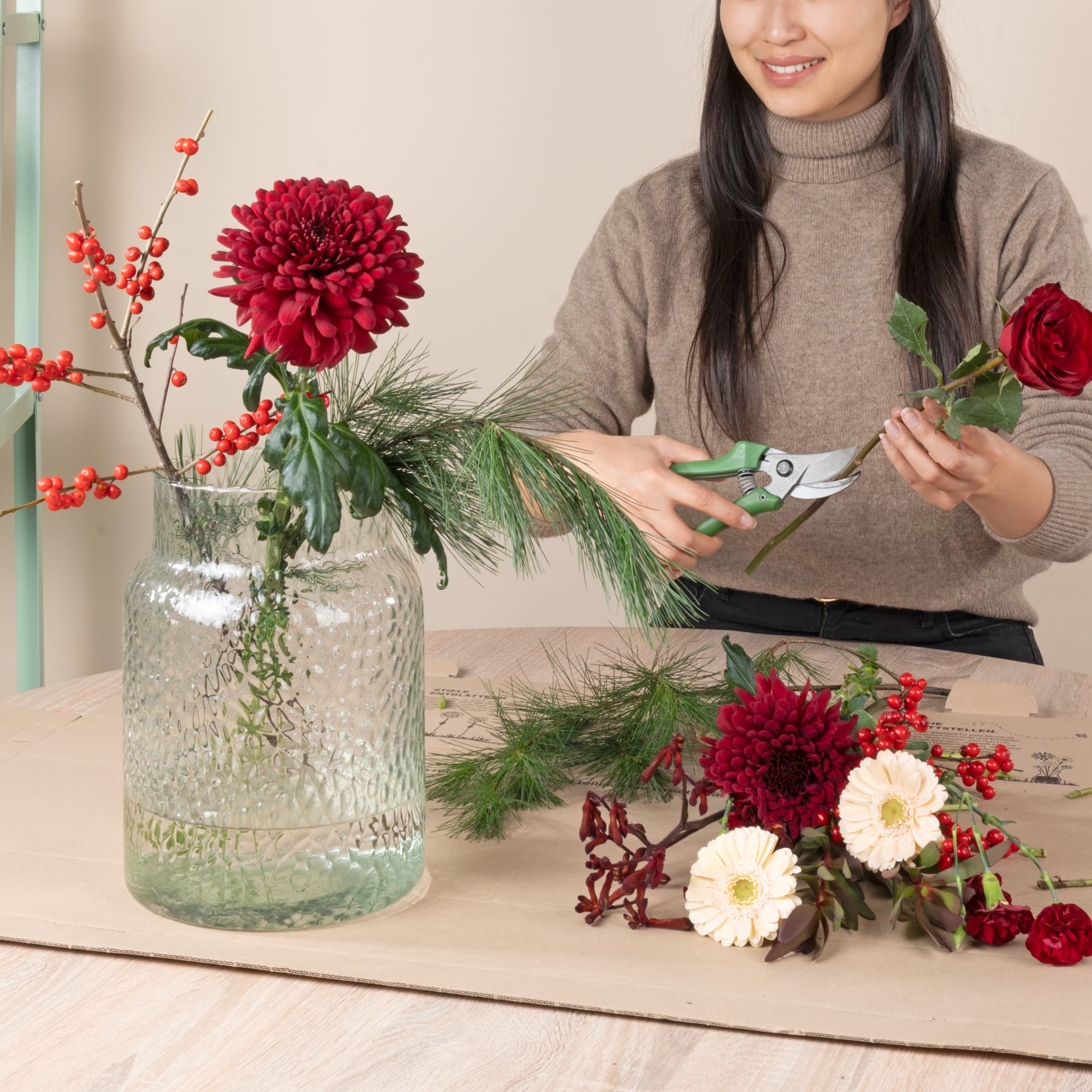 Eine Frau in einem beigen Pullover arrangiert eine Vase mit leuchtend roten und weißen Blumen, umgeben von festlicher Weihnachtsdekoration auf einem Holztisch.