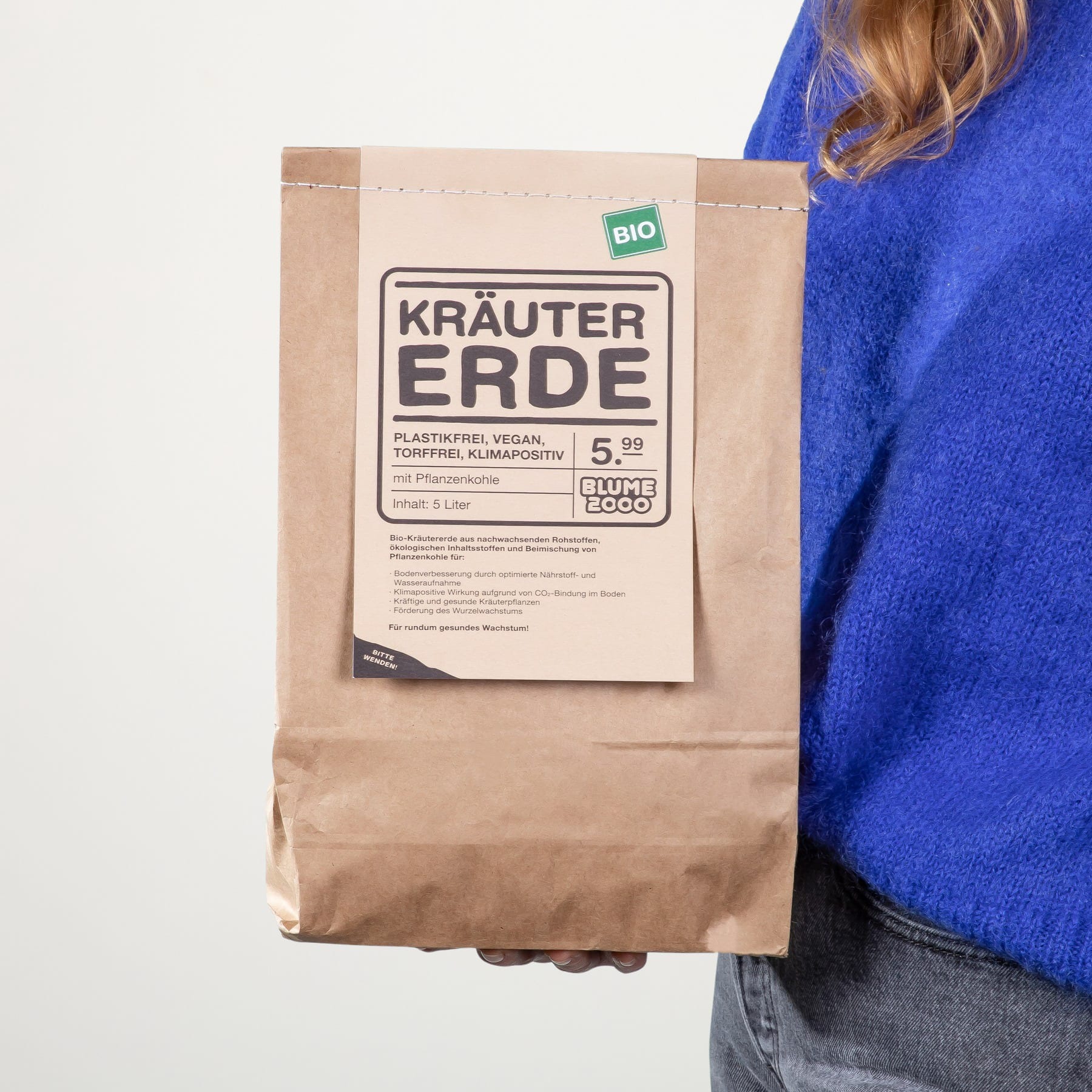 Die Hand einer Person hält vor weißem Hintergrund eine braune Papiertüte mit der Aufschrift „Krauter Erde“.