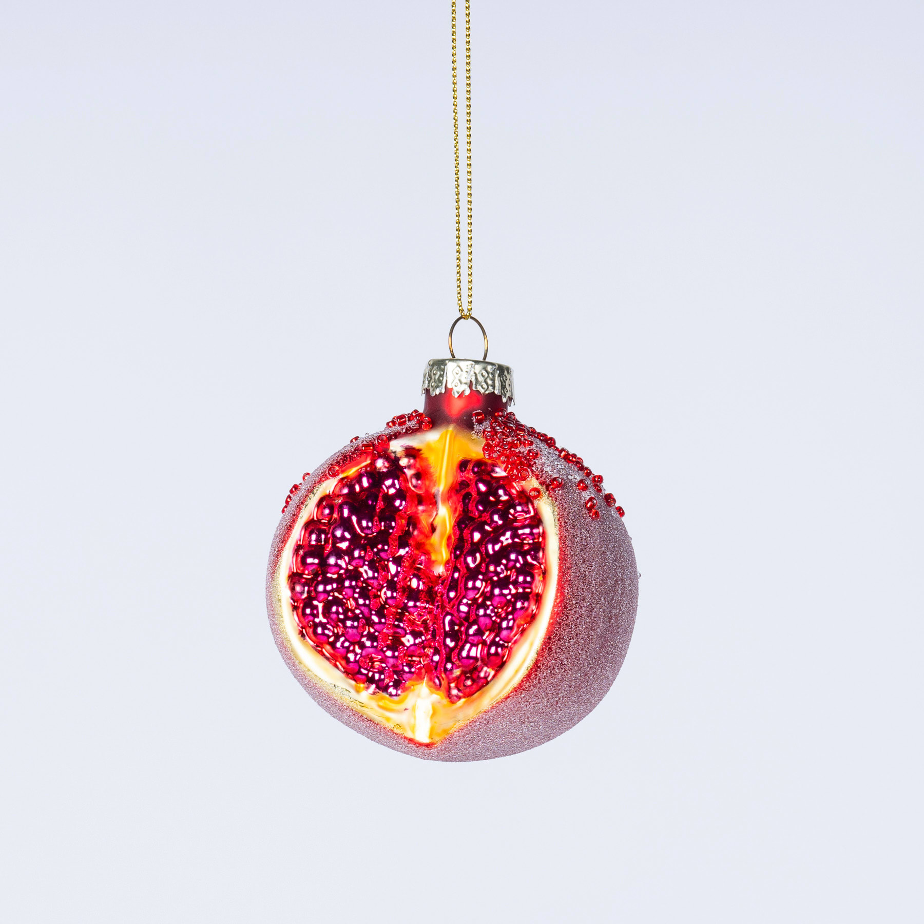 Vor einem schlichten weißen Hintergrund hängt ein Glasornament in Form eines Granatapfels, dessen leuchtend rote Kerne durch das transparente Glas sichtbar sind.