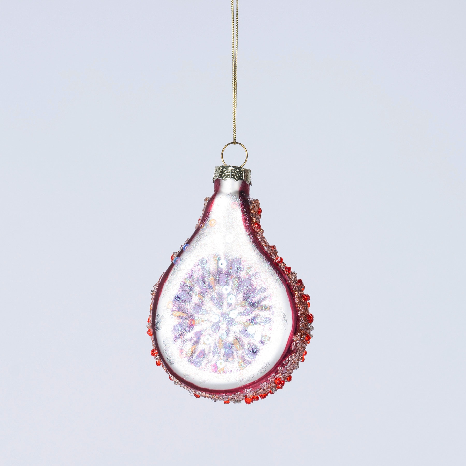 Ein zartes Glasornament in Form einer Drachenfrucht mit einer leuchtend roten Außenseite und einer funkelnden, kristallartigen Innenseite hängt vor einem schlichten, hellen Hintergrund.