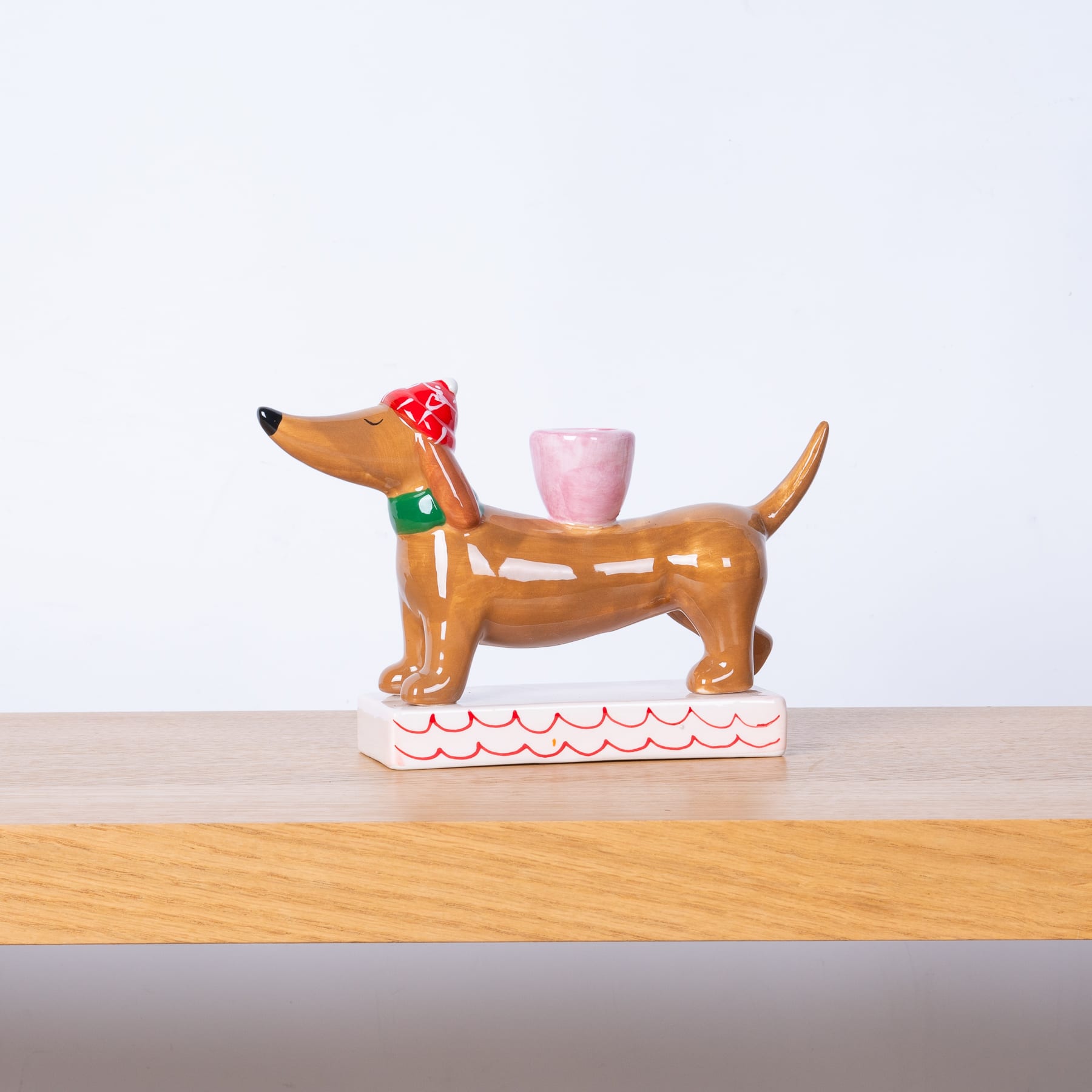 Eine farbenfrohe Keramikfigur eines Dackels mit einer roten Schleife, der auf einem weißen Sockel steht, daneben steht eine rosa Tasse, vor einem schlichten weißen Hintergrund.