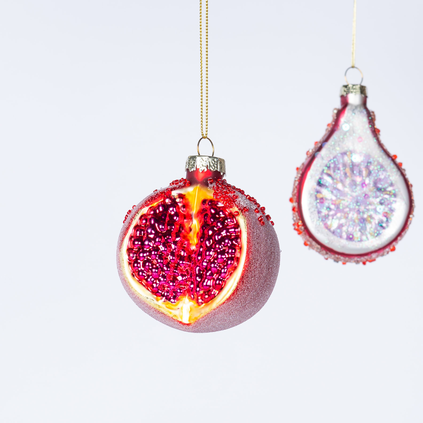 Vor einem schlichten weißen Hintergrund hängen zwei kunstvoll verzierte Weihnachtsornamente aus Glas, eines davon ein leuchtend roter Granatapfel und das andere eine funkelnde, schillernde Tropfenform.