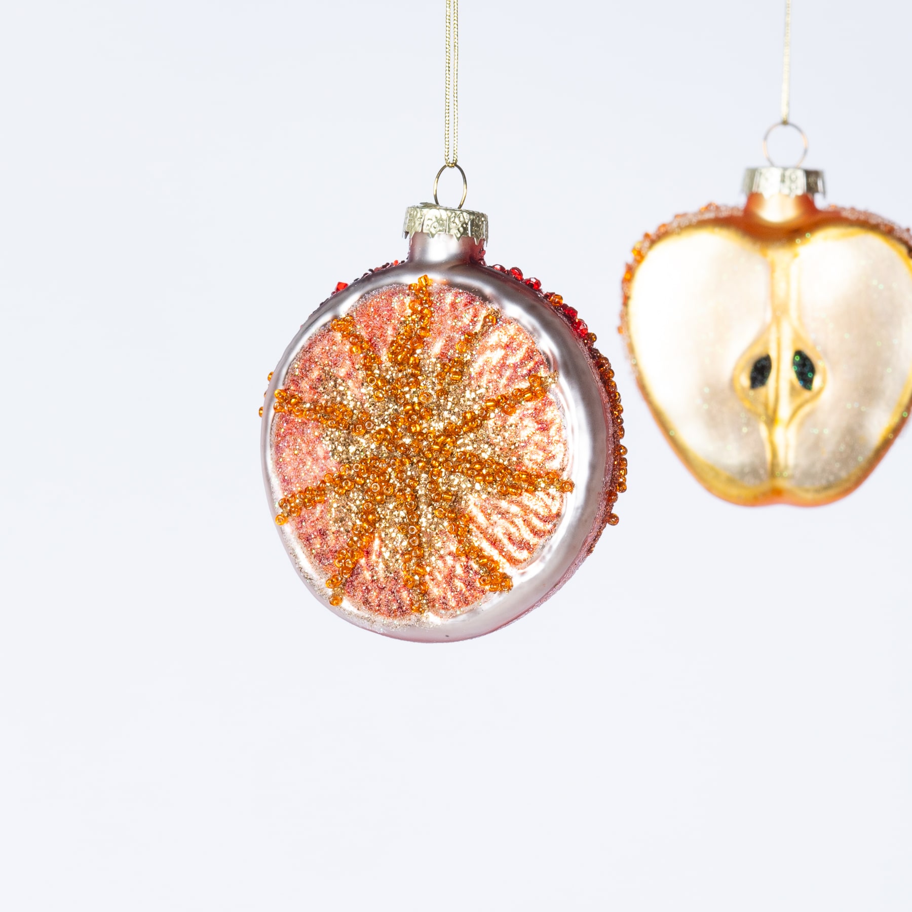 Das Bild zeigt zwei hängende Glasornamente auf weißem Hintergrund. Ein Ornament ist eine runde, rosafarbene Kugel mit einem glitzernden orangefarbenen Muster, das an ein Stern- oder Sonnenmuster erinnert. Das andere Ornament ist eine gelbe, birnenförmige Kugel mit einem schlichten, cartoonhaften Gesicht darauf.