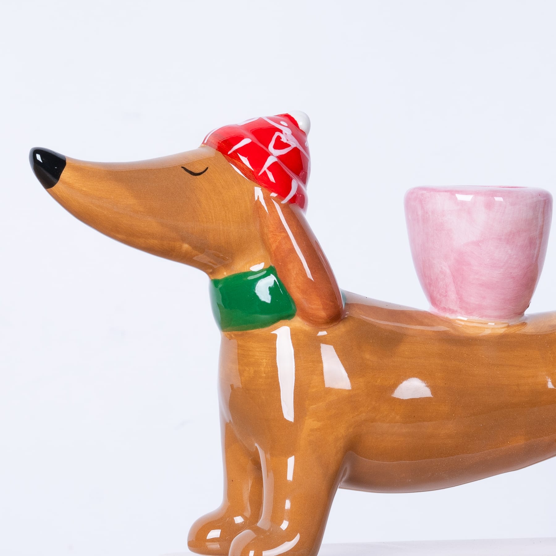 Eine Keramikfigur eines Hundes mit einer roten Schleife, der neben einer rosa Keramiktasse auf weißem Hintergrund sitzt.