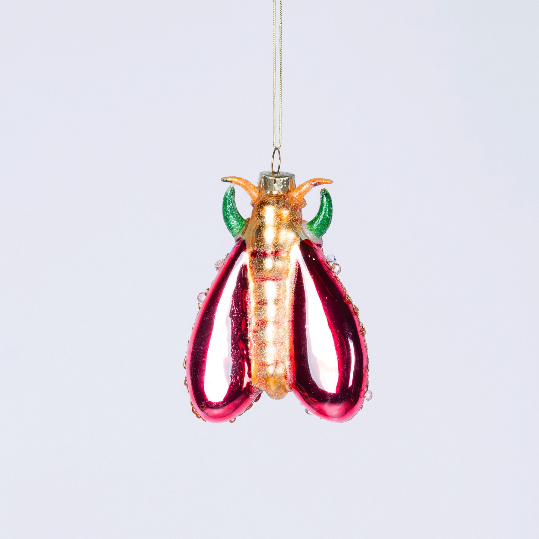 Ein farbenfrohes und skurriles Glasornament in Form eines Hummers oder einer Krabbe, mit einem metallisch goldenen Körper und leuchtend roten und grünen Anhängseln, das vor einem schlichten weißen Hintergrund schwebt.