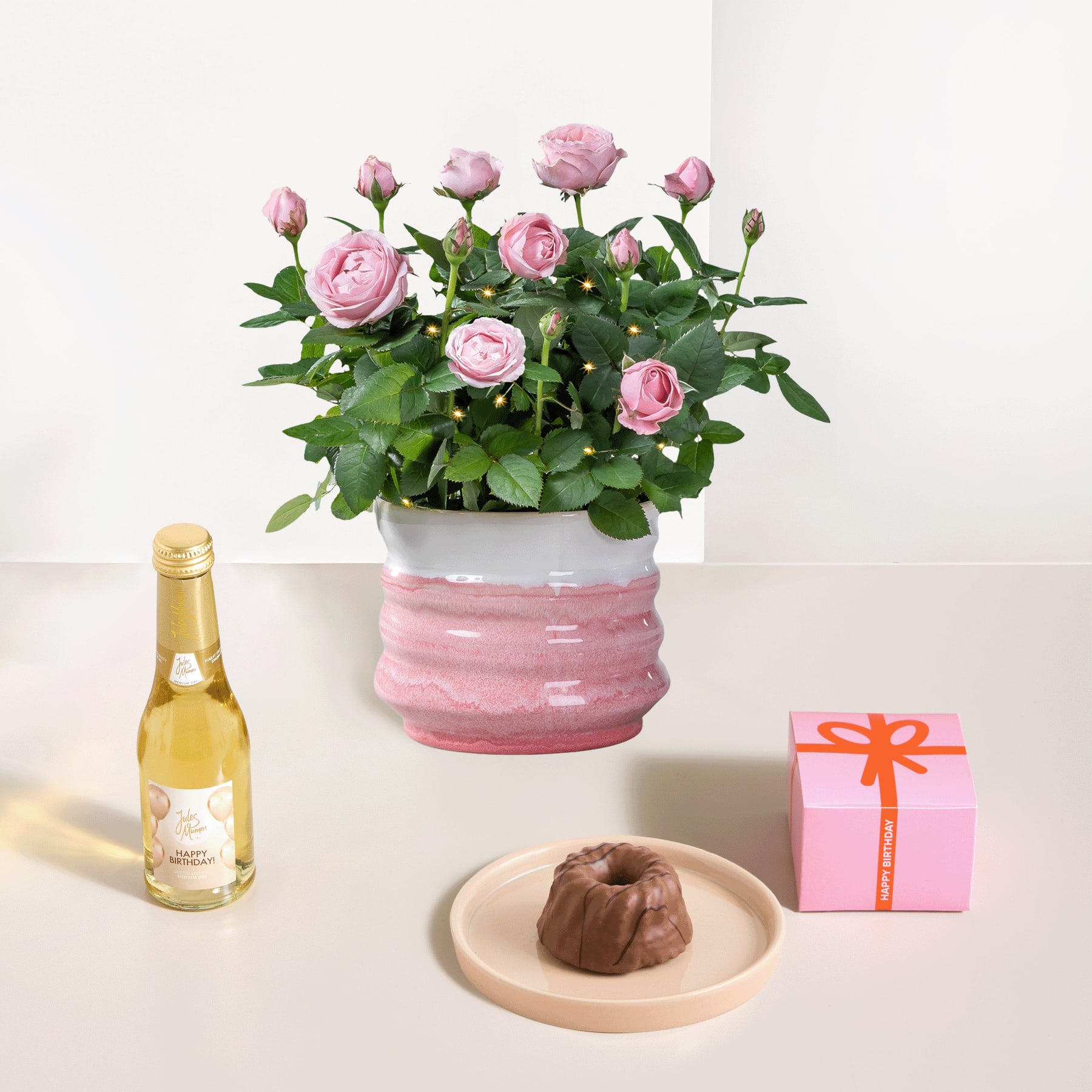 Ein Strauß zartrosa Rosen steht in einem rosa Keramiktopf, dazu gibt es eine Flasche Weißwein und ein kleines Dessert auf einem Teller, alles auf einem schlichten weißen Hintergrund arrangiert.