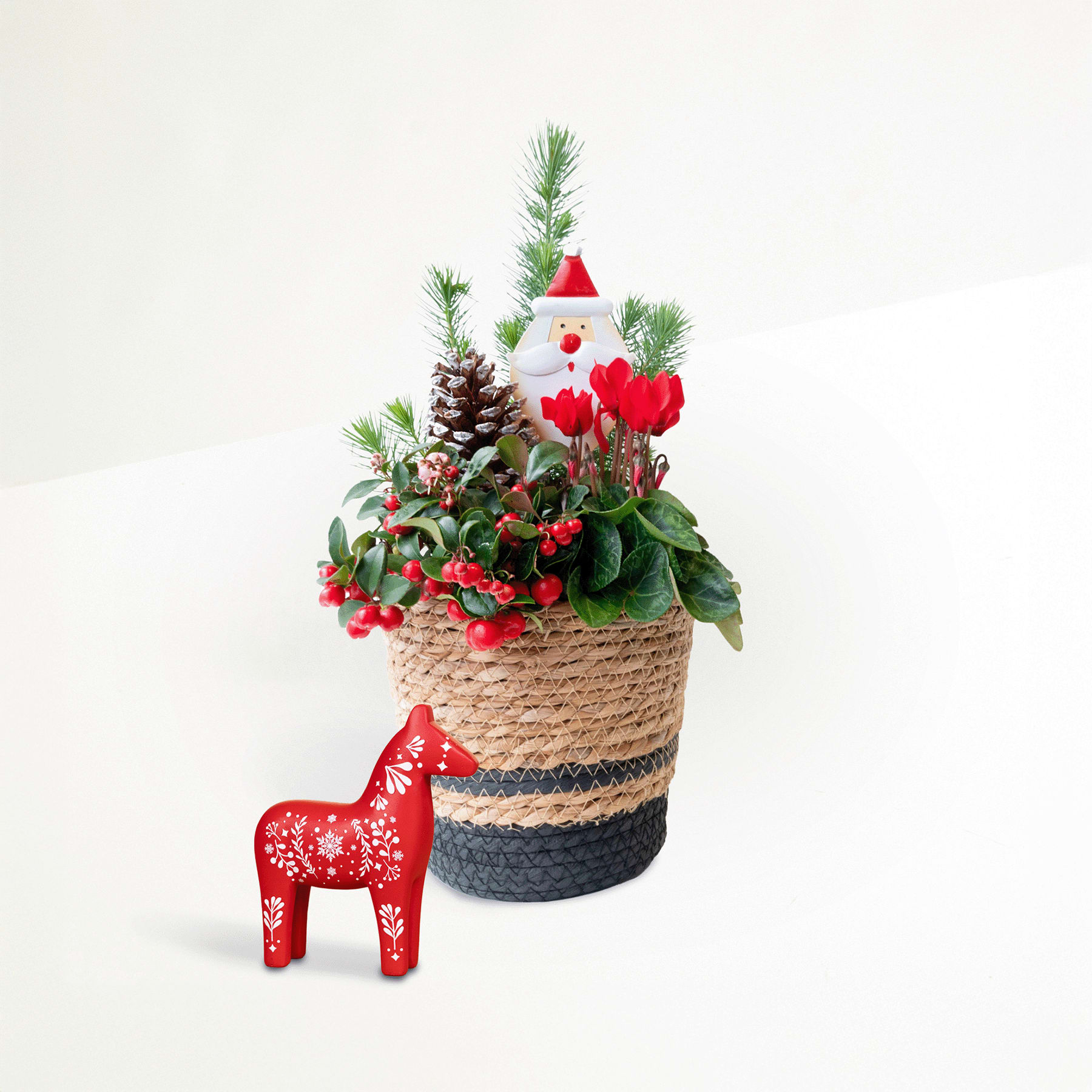 Ein festliches Weihnachtsarrangement mit einer Weihnachtsmannfigur, Tannenzweigen, roten Tulpen und einem dekorativen Holzpferd, alles in einem geflochtenen Korb vor einem schlichten weißen Hintergrund.