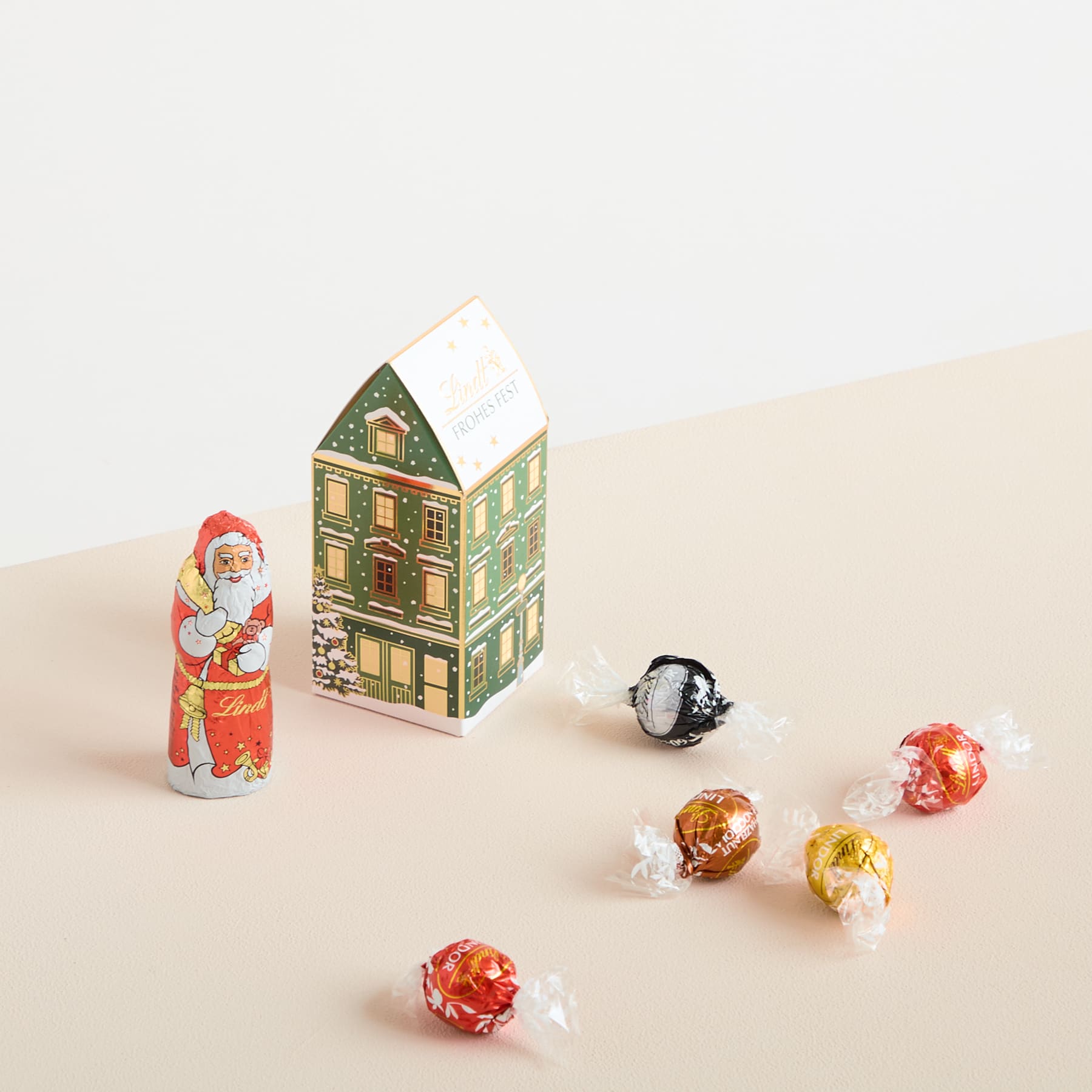 Eine festliche Szene mit einer kleinen Weihnachtsmannfigur, einem dekorativen hausförmigen Behälter und verschiedenen verpackten Süßigkeiten, die auf einer hellen Oberfläche verstreut sind.