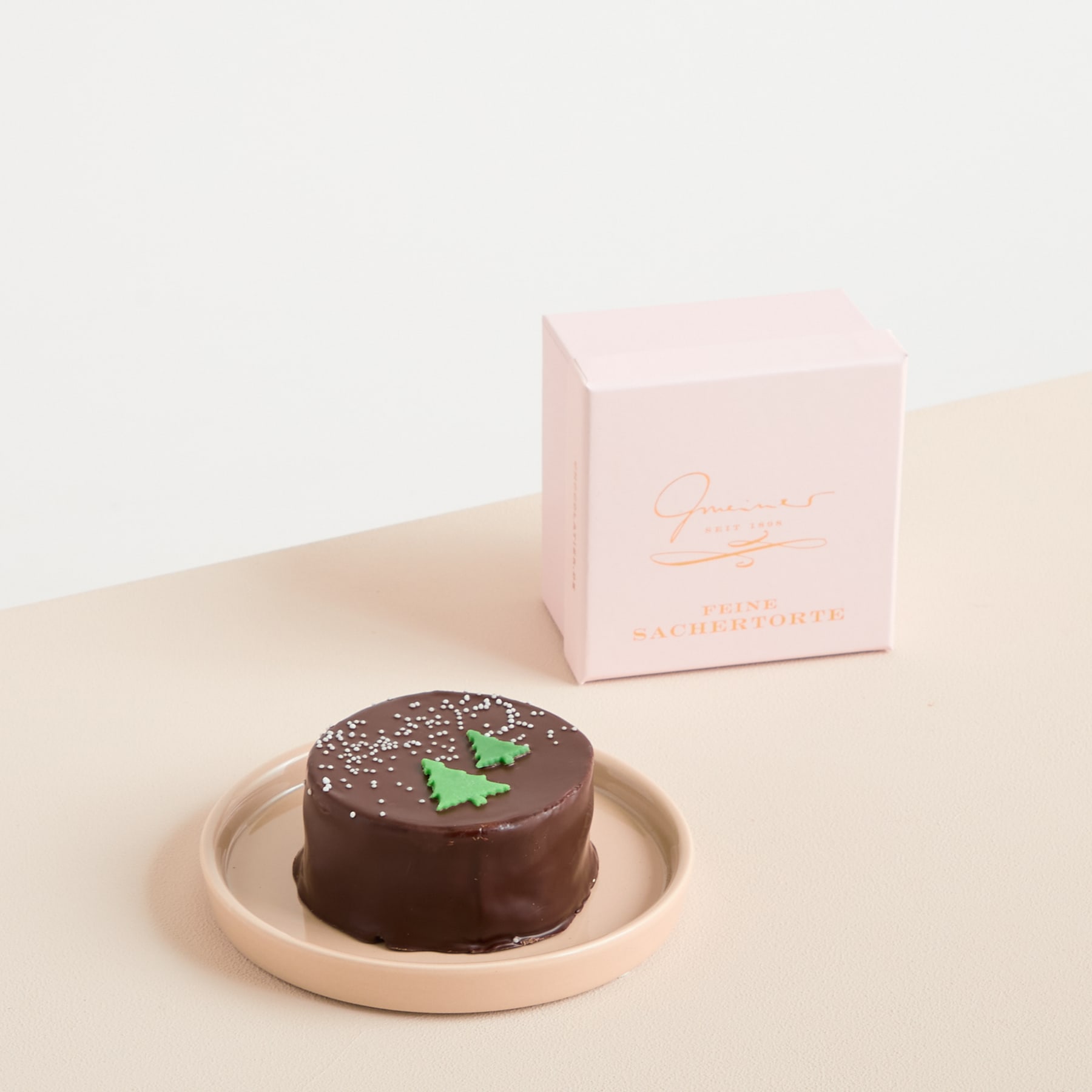 Ein kleines, rundes Schokoladendessert liegt auf einem blassrosa Teller, obenauf ist ein grünes Blattdekor angebracht. Im Hintergrund ist eine rosa Schachtel mit weißem Text zu sehen.