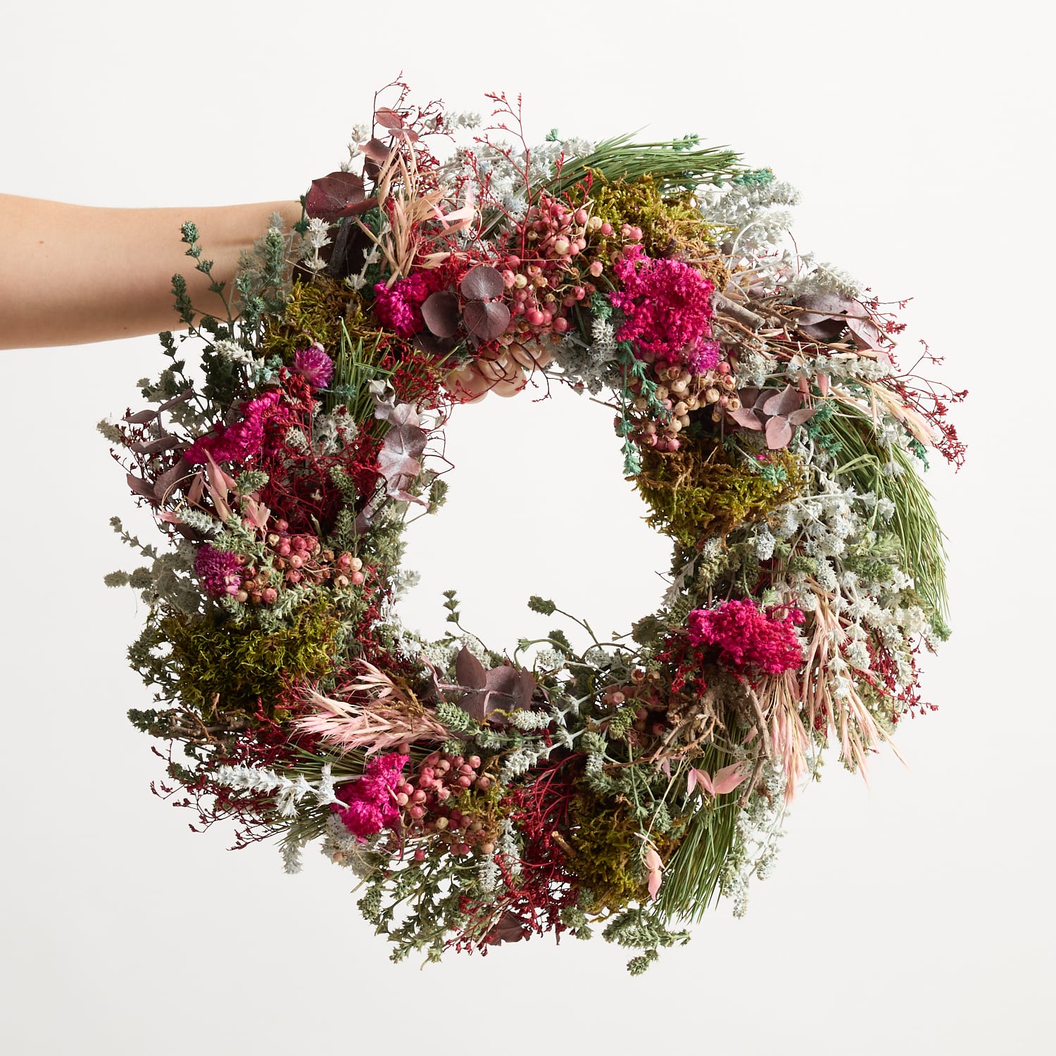 Ein lebendiger, üppiger Kranz aus verschiedenen Blumen, Blättern und Naturelementen wird vor einem schlichten weißen Hintergrund in den Händen einer Person gehalten.