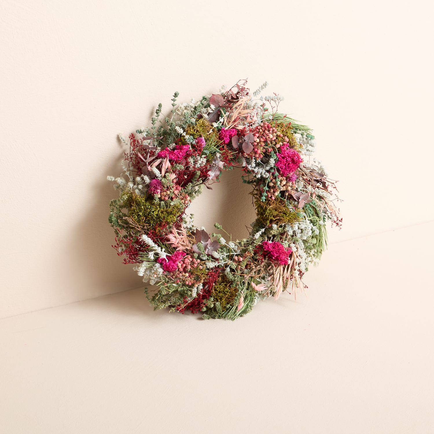 Ein wunderschöner, üppiger Kranz aus verschiedenen Blumen, Grünpflanzen und getrockneten Elementen, der an einer schlichten, hellen Wand hängt.