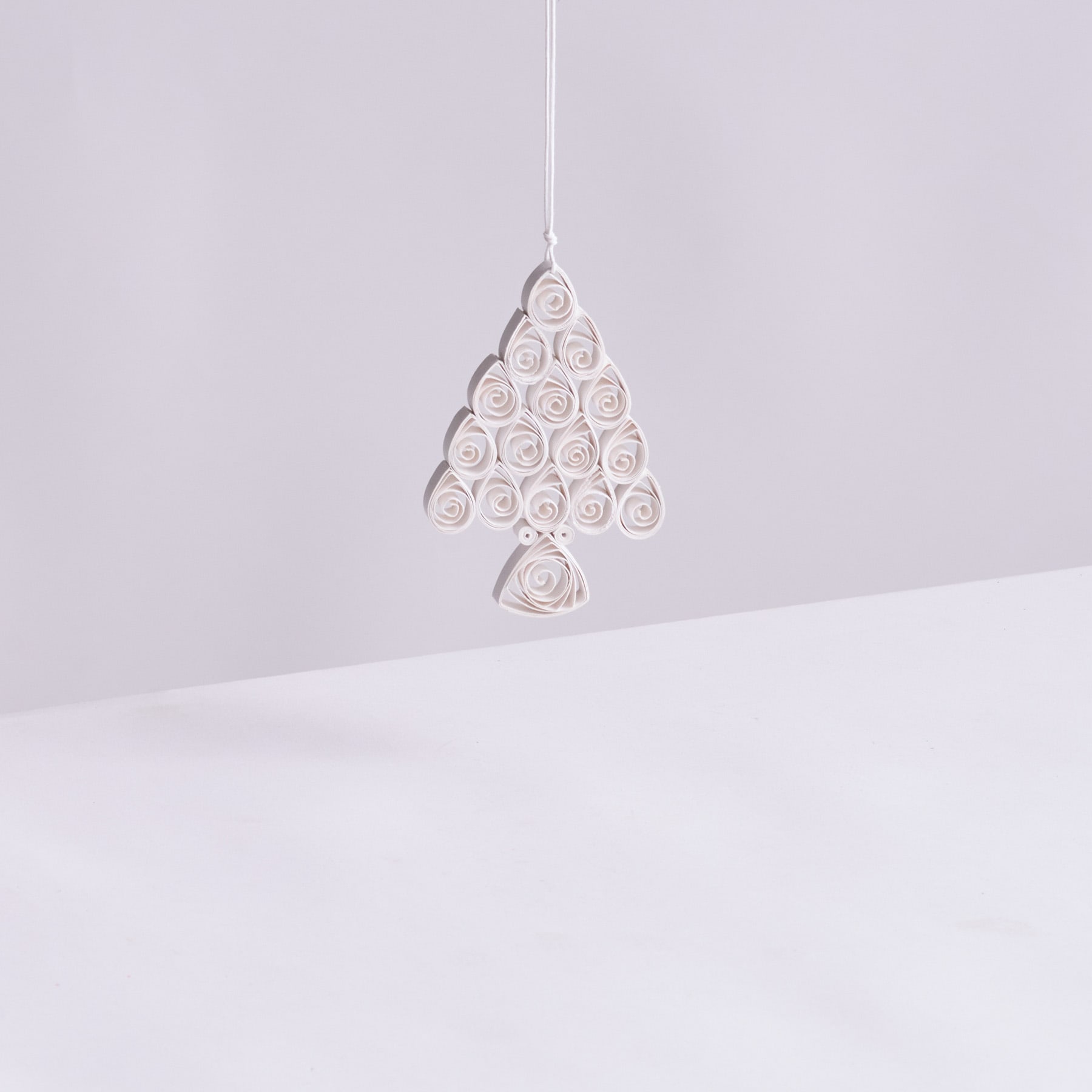 Ein zartes, weißes Papierornament in Form eines Weihnachtsbaums hängt vor einem schlichten, hellen Hintergrund.