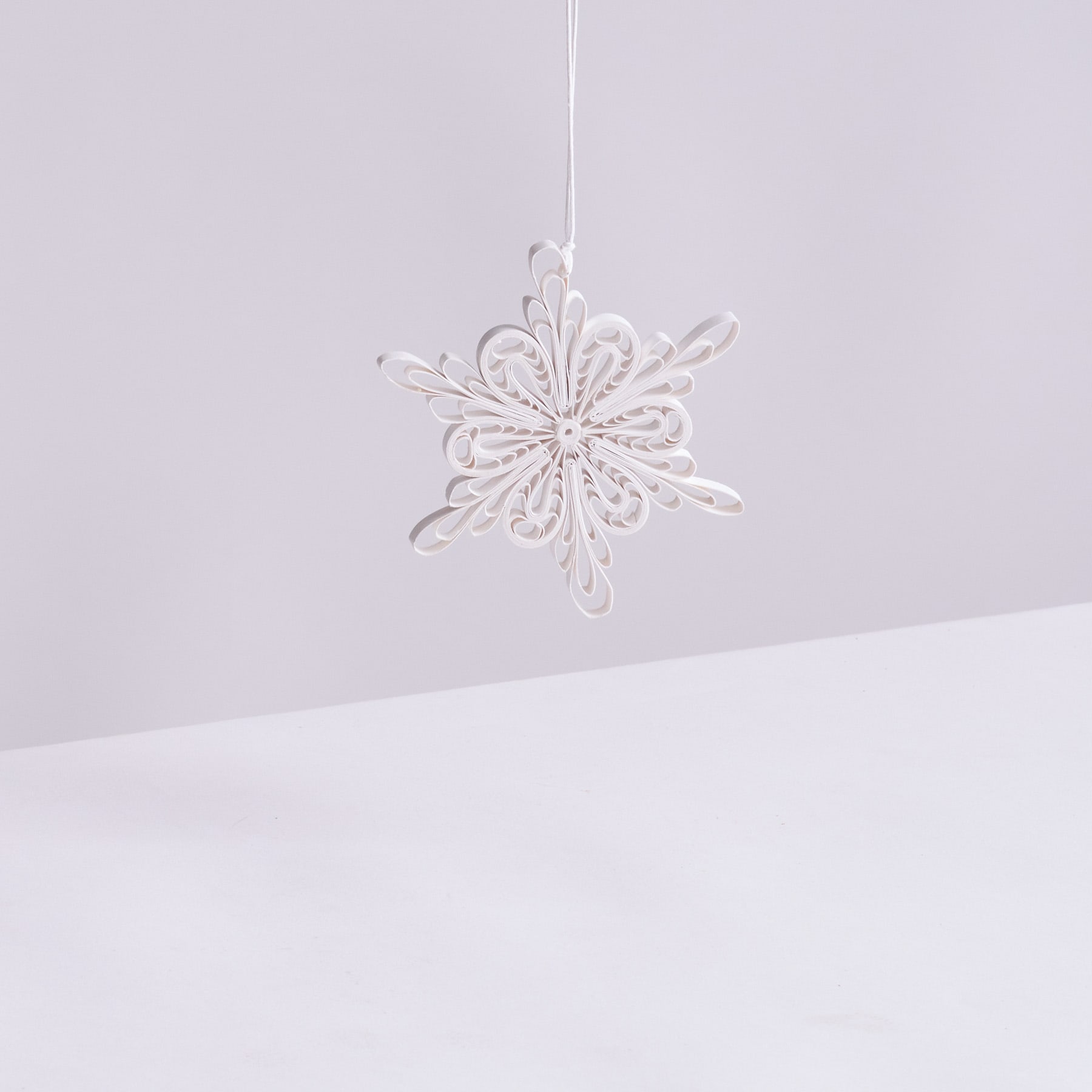 Ein zartes, spitzenartiges Schneeflockenornament hängt vor einem schlichten, blassen Hintergrund.