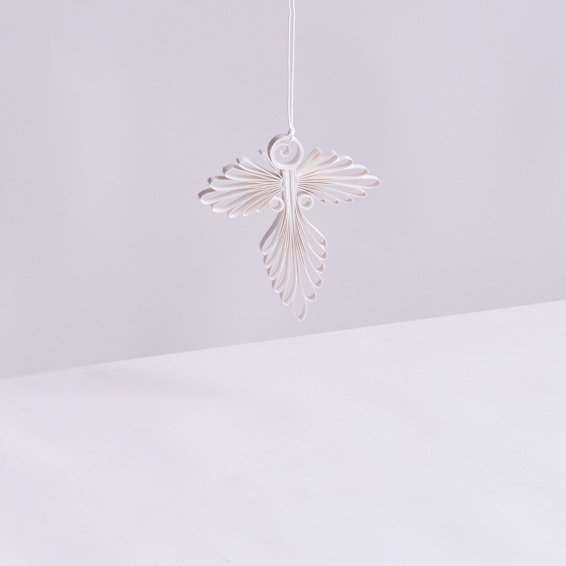 Ein zartes, weißes, kreuzförmiges Ornament hängt vor einem schlichten, blassen Hintergrund.