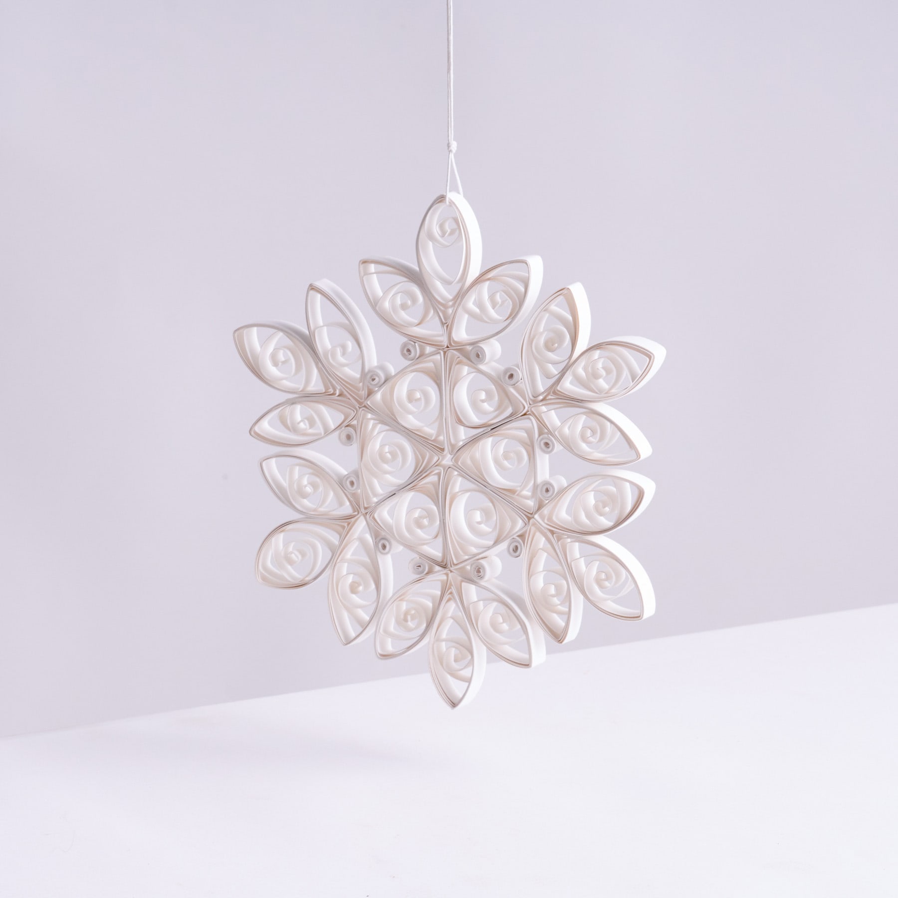 Ein zartes, kompliziertes, schneeflockenartiges Ornament aus weißem, durchscheinendem Material hängt vor einem schlichten, hellen Hintergrund.