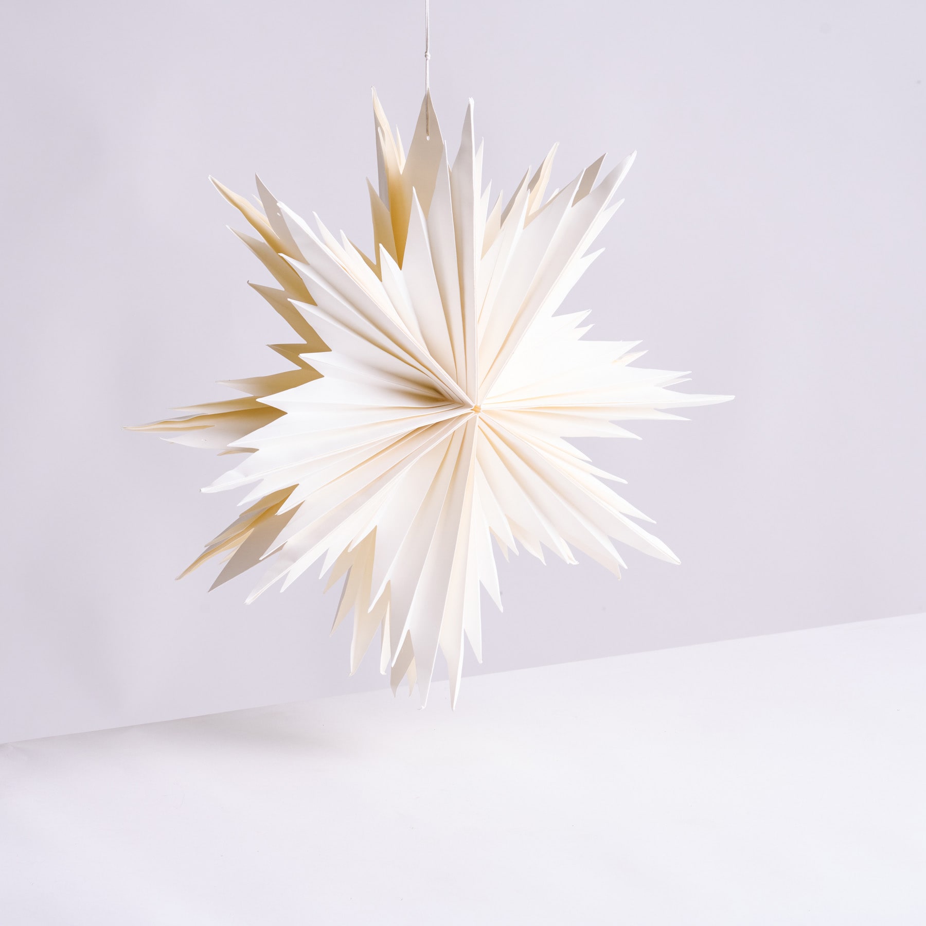Eine zarte, sternförmige Dekoration aus weißem Papier mit scharfen, eckigen Spitzen schwebt vor einem schlichten, hellen Hintergrund.