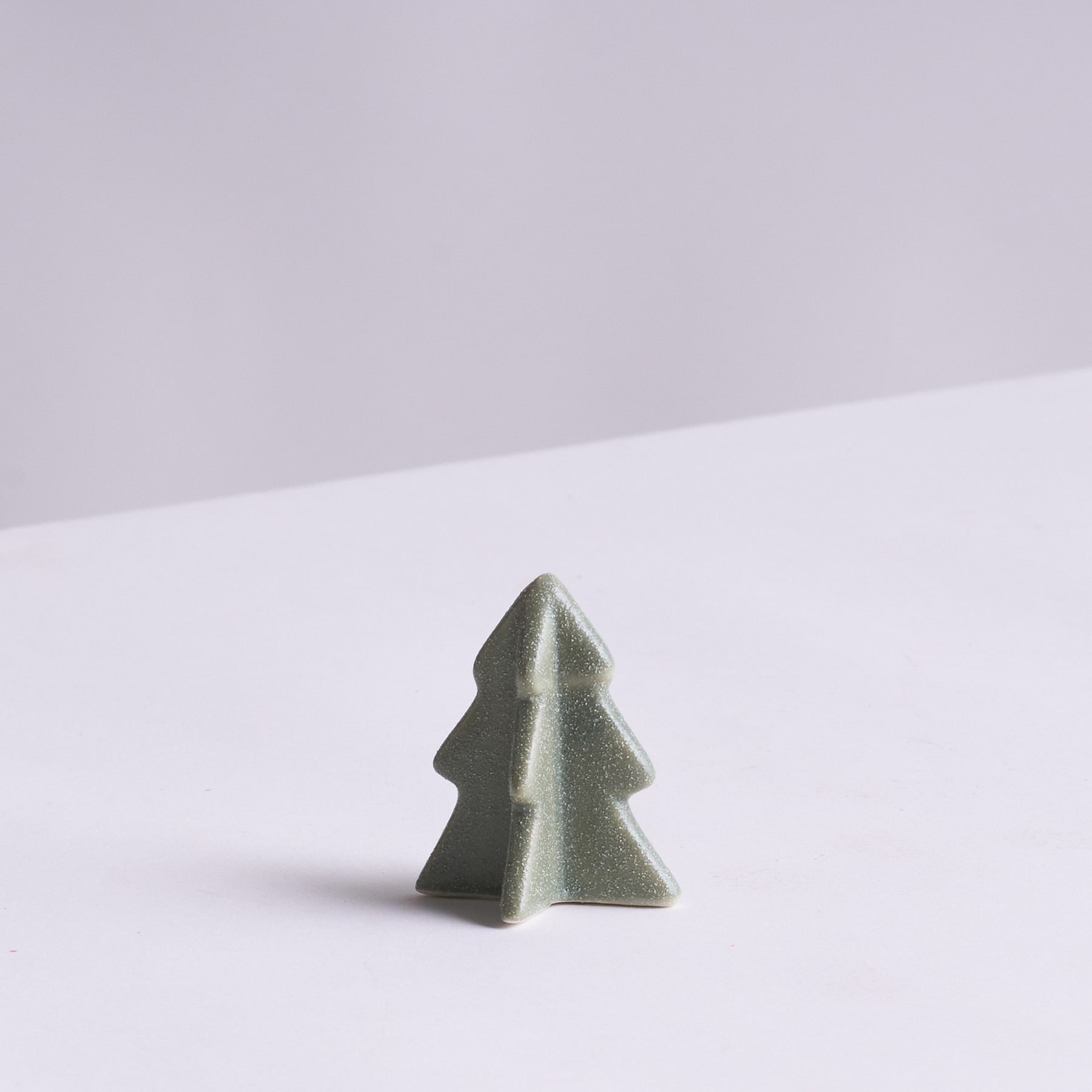 Ein kleines, grünes, dreieckiges Objekt, das einem Weihnachtsbaum ähnelt, steht allein vor einem schlichten, hellen Hintergrund.