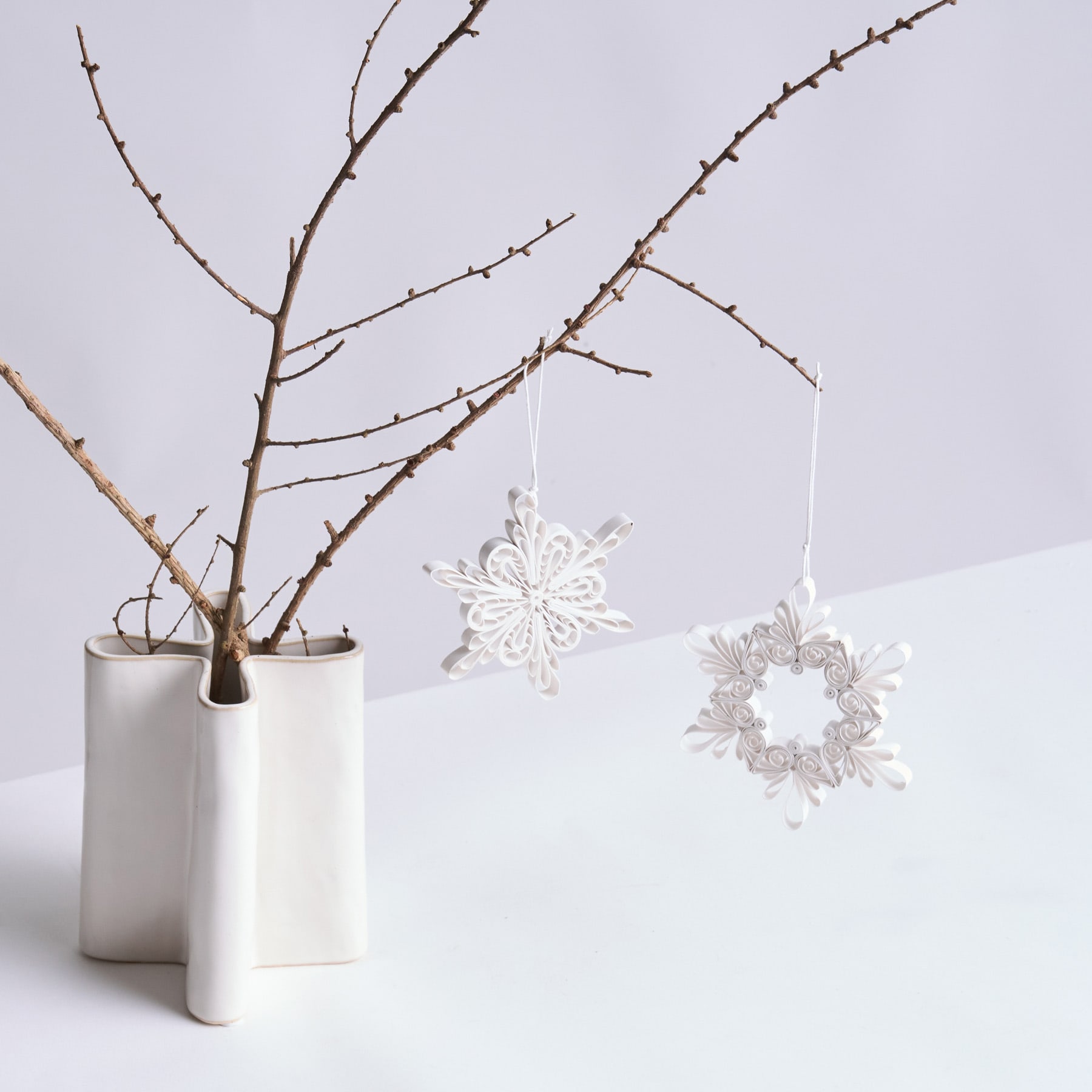 Eine weiße Keramikvase enthält kahle Äste, an denen vor einem schlichten weißen Hintergrund zarte weiße Schneeflockenornamente hängen.