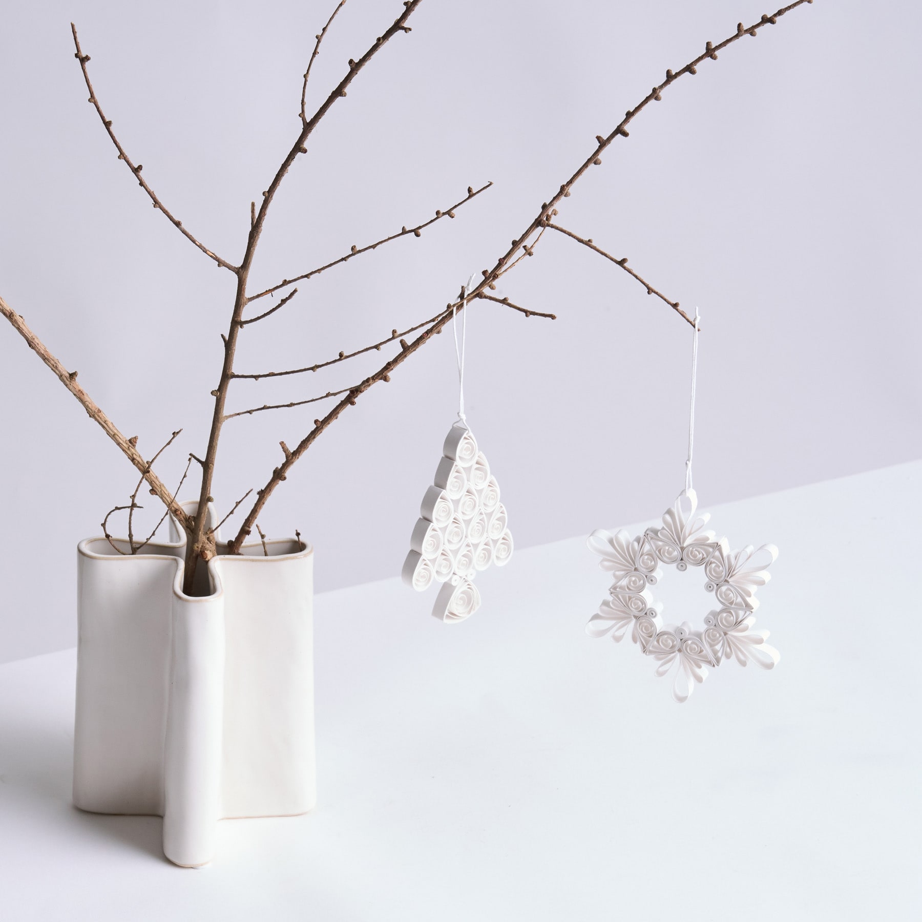 Eine minimalistische Winterszene mit einer Vase mit kahlen Zweigen, einem zarten weißen Ornament und einer kranzartigen Dekoration, die vor einem schlichten weißen Hintergrund hängt.