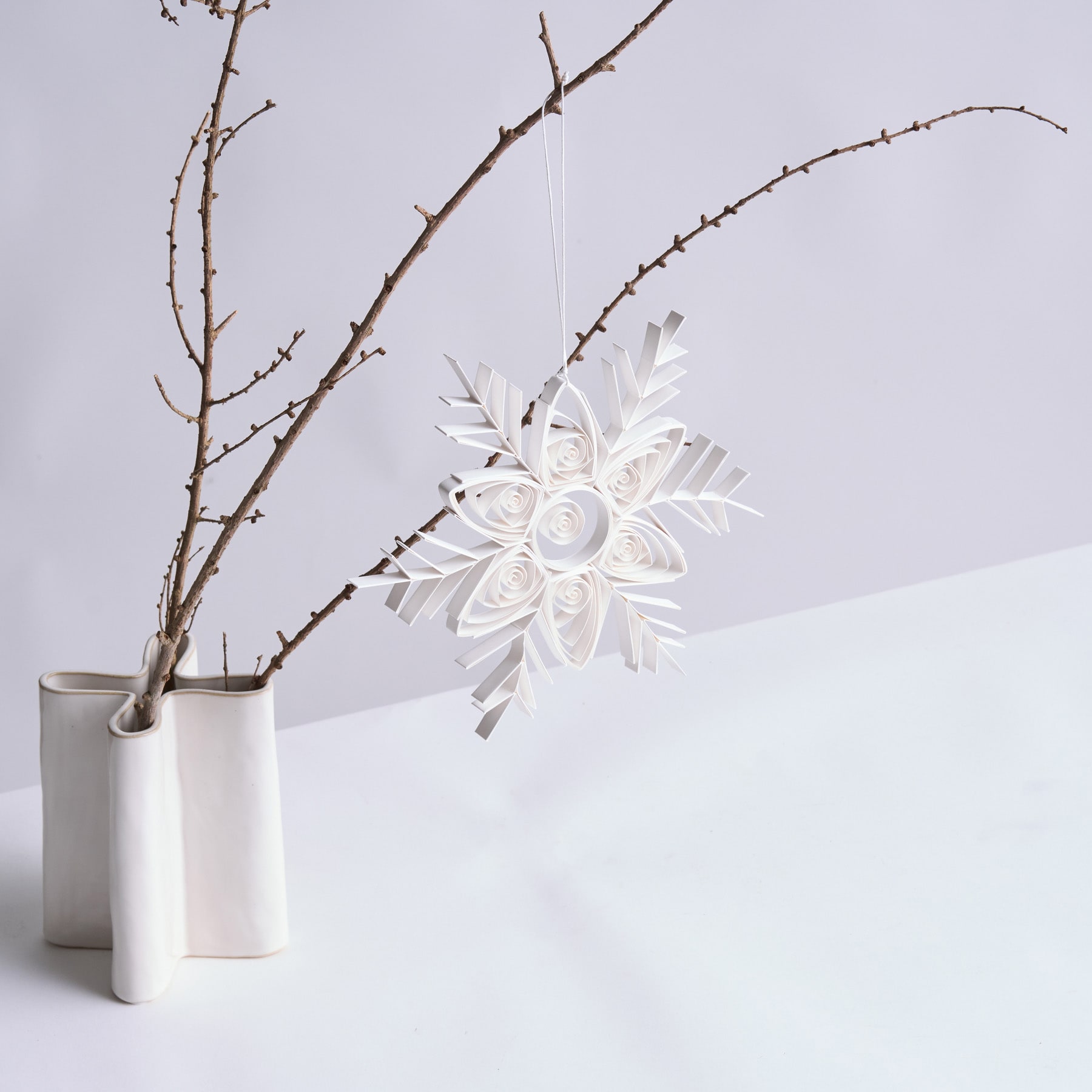 Vor einem schlichten weißen Hintergrund hängt ein zartes Schneeflockenornament aus weißem Papier an einer Vase voller kahler Äste.