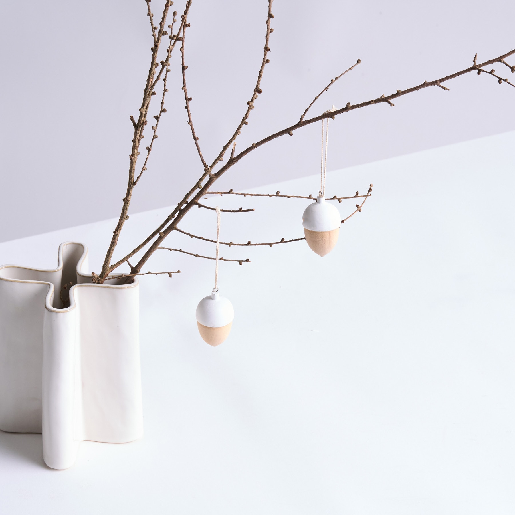 Ein minimalistisches Arrangement mit einer Vase mit kahlen Zweigen und daran hängenden zarten weißen Eiern vor einem schlichten weißen Hintergrund.