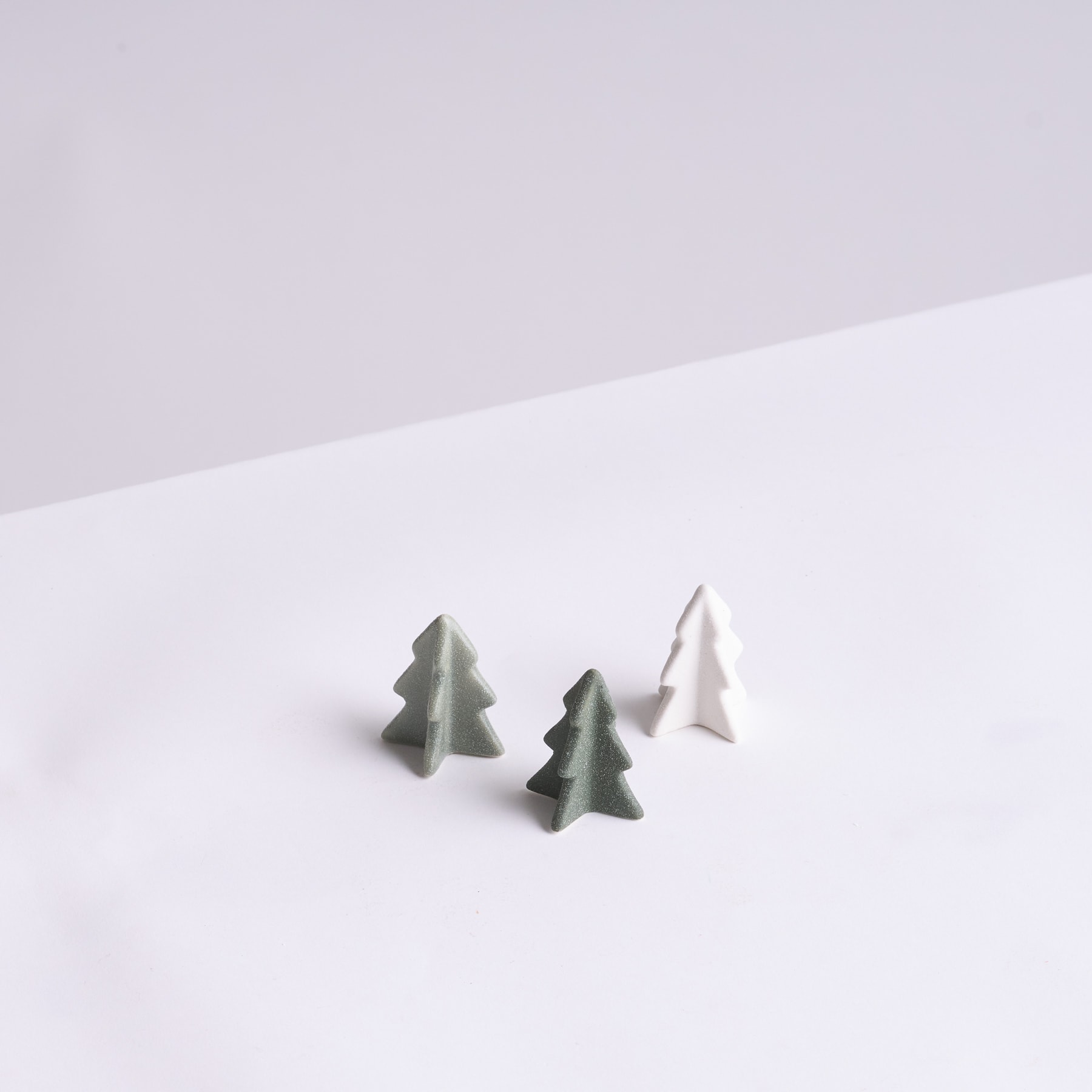 Auf einem schlichten weißen Untergrund sitzen drei kleine, stilisierte Christbaumfiguren aus Stein oder Keramik.