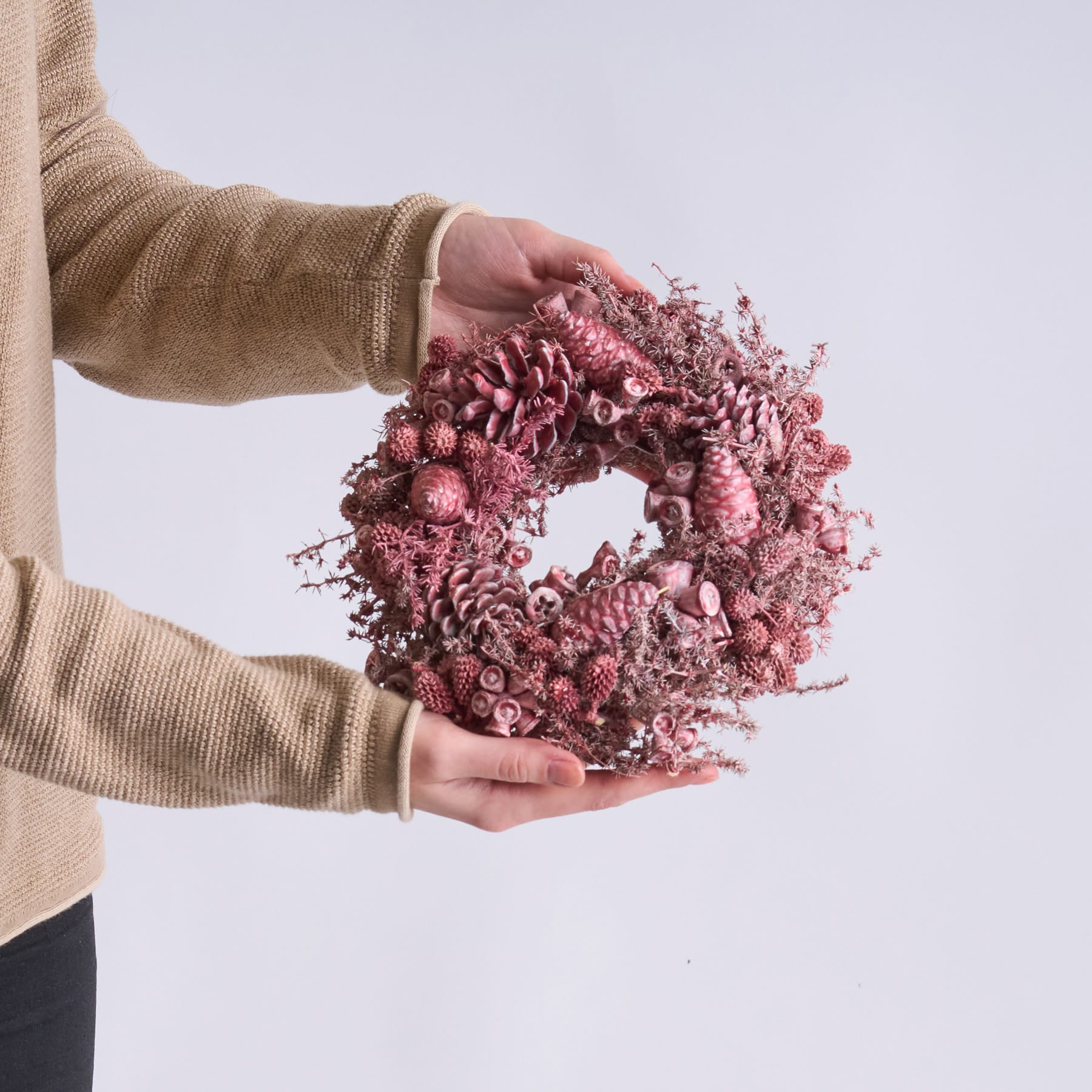 Die Hände einer Person halten einen leuchtend rosafarbenen Kranz aus getrockneten Blumen und Tannenzapfen vor einem schlichten, hellen Hintergrund.