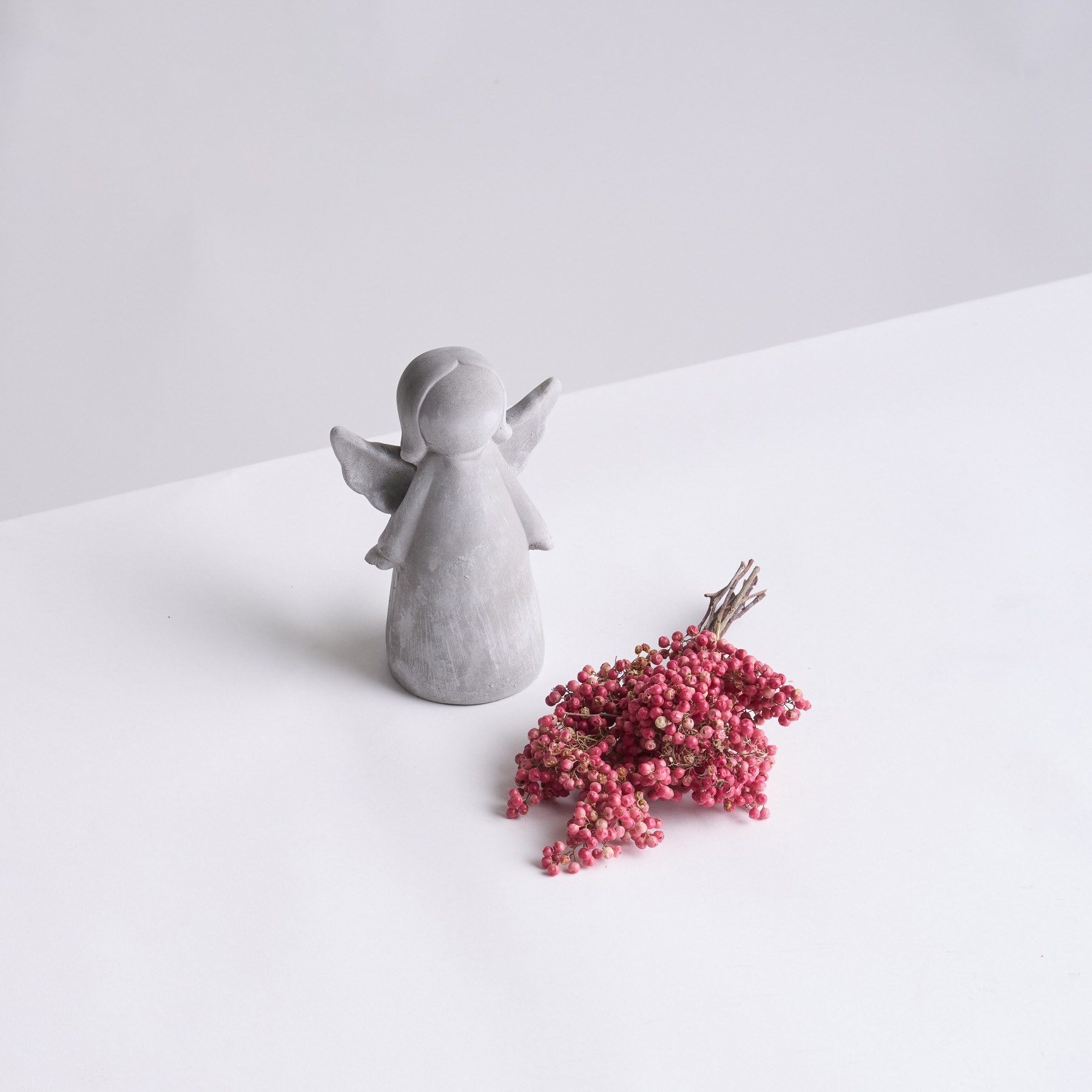 Eine kleine, graue Engelsfigur aus Keramik steht neben einem verstreuten Haufen leuchtend roter Beeren auf einem schlichten weißen Hintergrund.