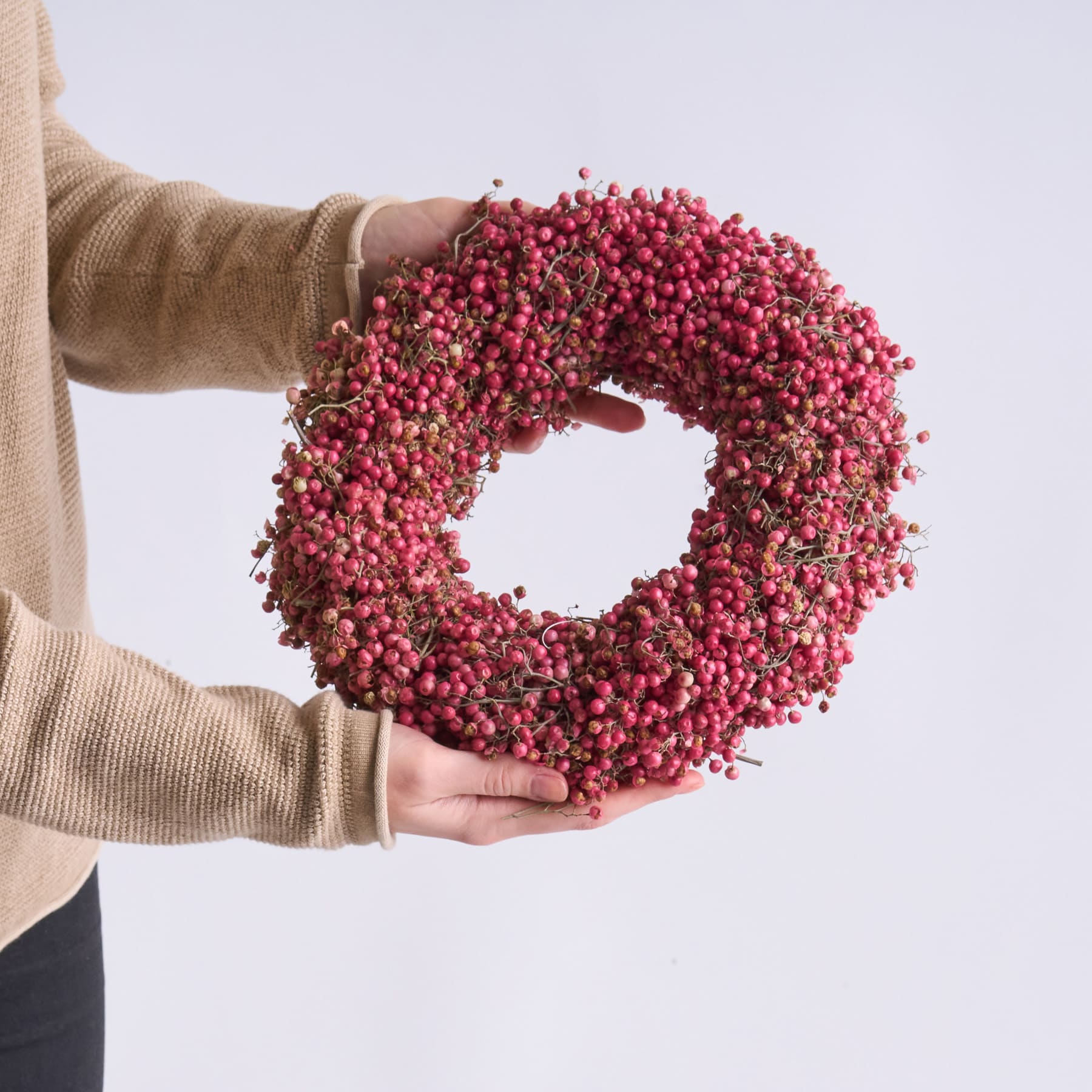 Vor einem schlichten weißen Hintergrund hält eine Person einen Kranz aus leuchtend roten Beeren in den Händen.
