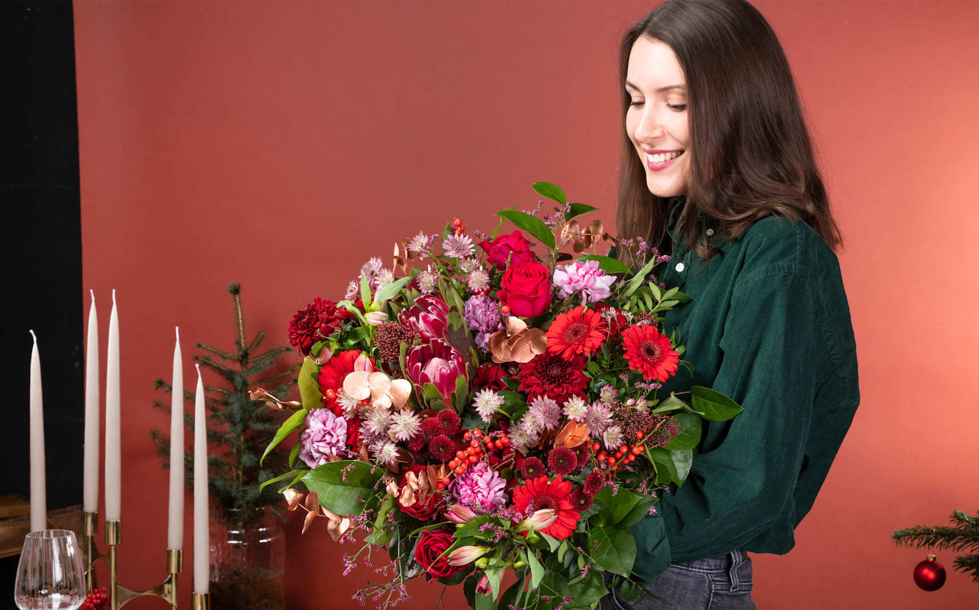 Eine junge Frau mit langem, dunklem Haar hält vor einem tiefroten Hintergrund einen großen, leuchtenden Strauß bunter Blumen, darunter rote, rosa und weiße Blüten.