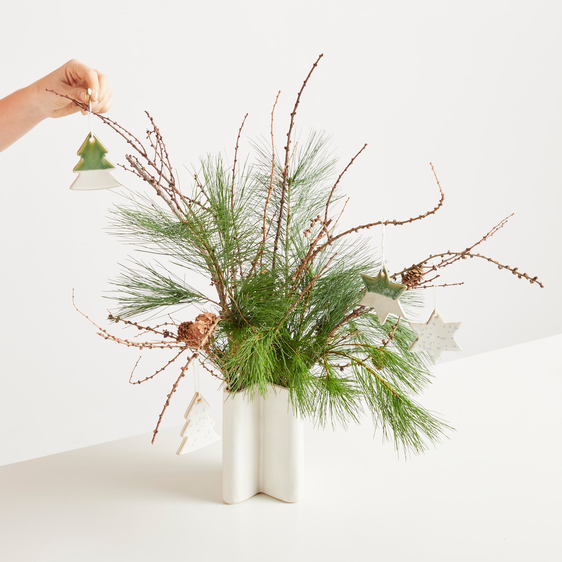 Eine Vase mit einem üppigen Arrangement aus Tannenzweigen, Zweigen und Grünpflanzen vor einem schlichten weißen Hintergrund.