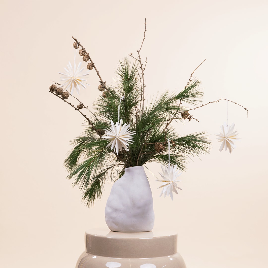 Eine weiße Keramikvase mit einem farbenfrohen Arrangement aus Tannenzweigen, getrockneten Blumen und weißen Gänseblümchen steht auf einem beigen Sockel vor einem schlichten, hellen Hintergrund.