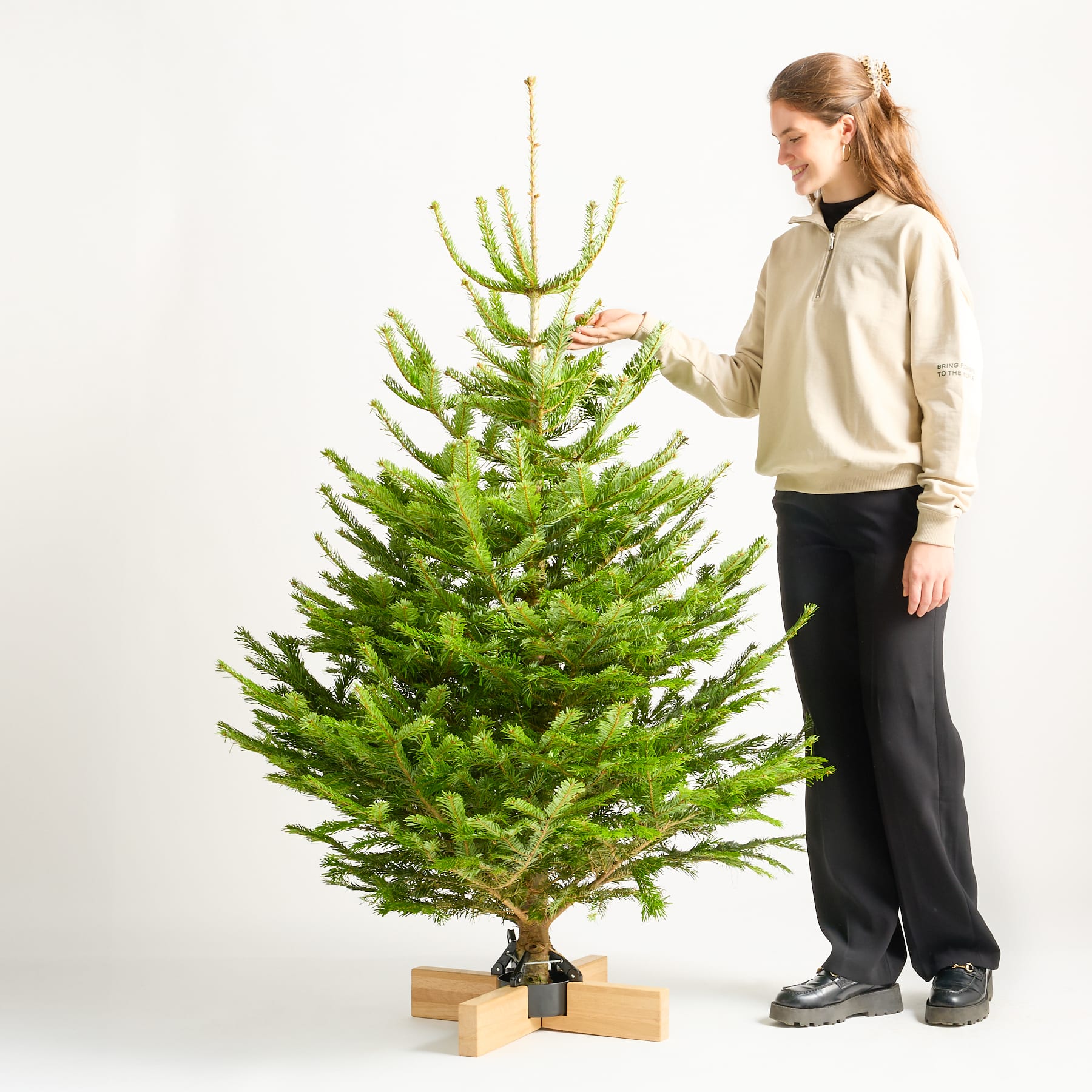 Eine junge Frau steht vor einem schlichten weißen Hintergrund neben einem großen, üppig grünen Weihnachtsbaum, der mit einem einfachen Stern als Spitze geschmückt ist.