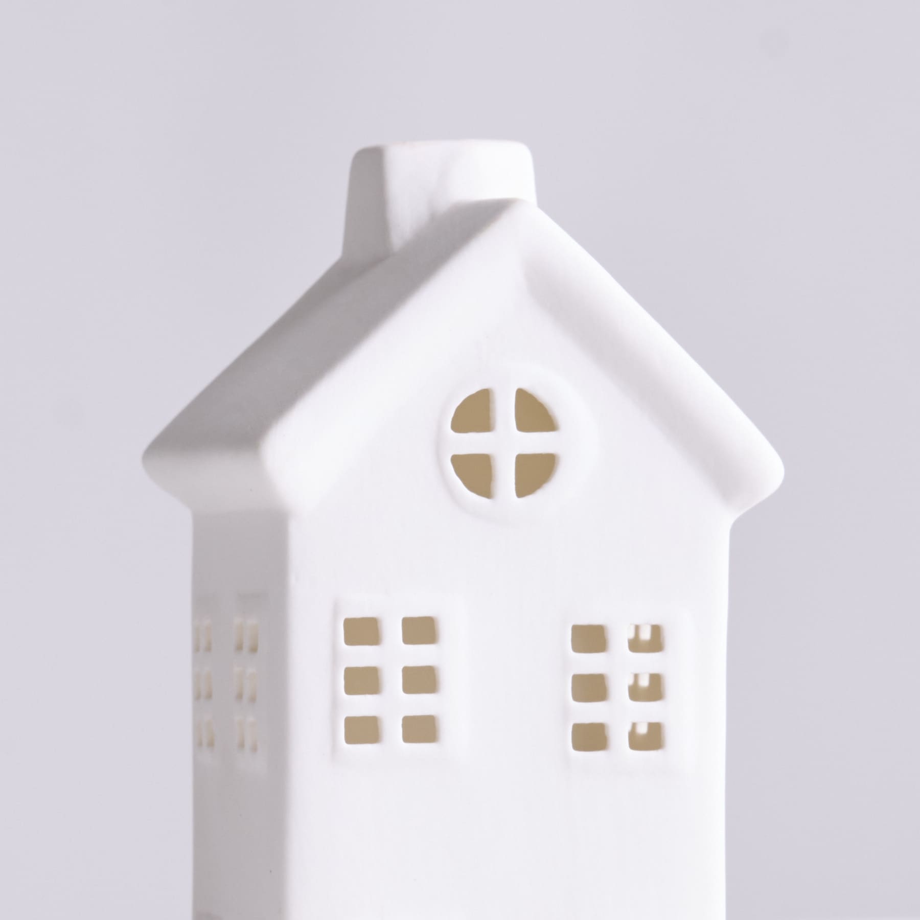 Vor einem schlichten, hellen Hintergrund steht ein kleines, weißes Keramikornament in Form eines Hauses mit Fenstern und einer Tür.